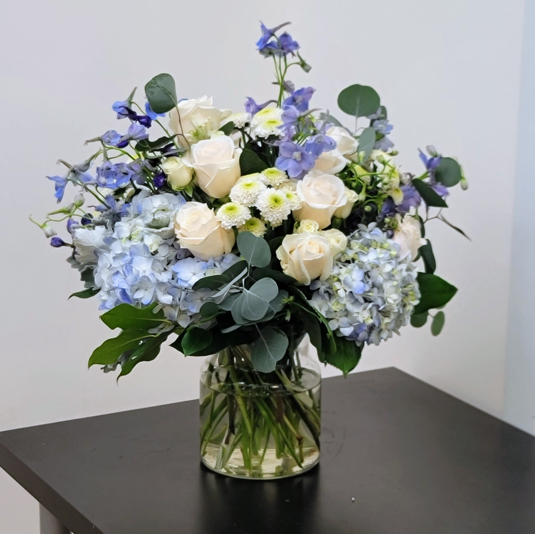 Clear Skies Bouquet - Bring a sense of calm and elegance with this serene arrangement of blue hydrangeas, soft white roses, and delicate light blue accents.   Designed to capture the beauty of clear skies, this bouquet is perfect for celebrations, a new baby, a celebration of life, or simply brightening any space. Expertly arranged in a classic clear glass vase.  Approximate Size: 18" H x 16" W. Order now for a touch of tranquility!  