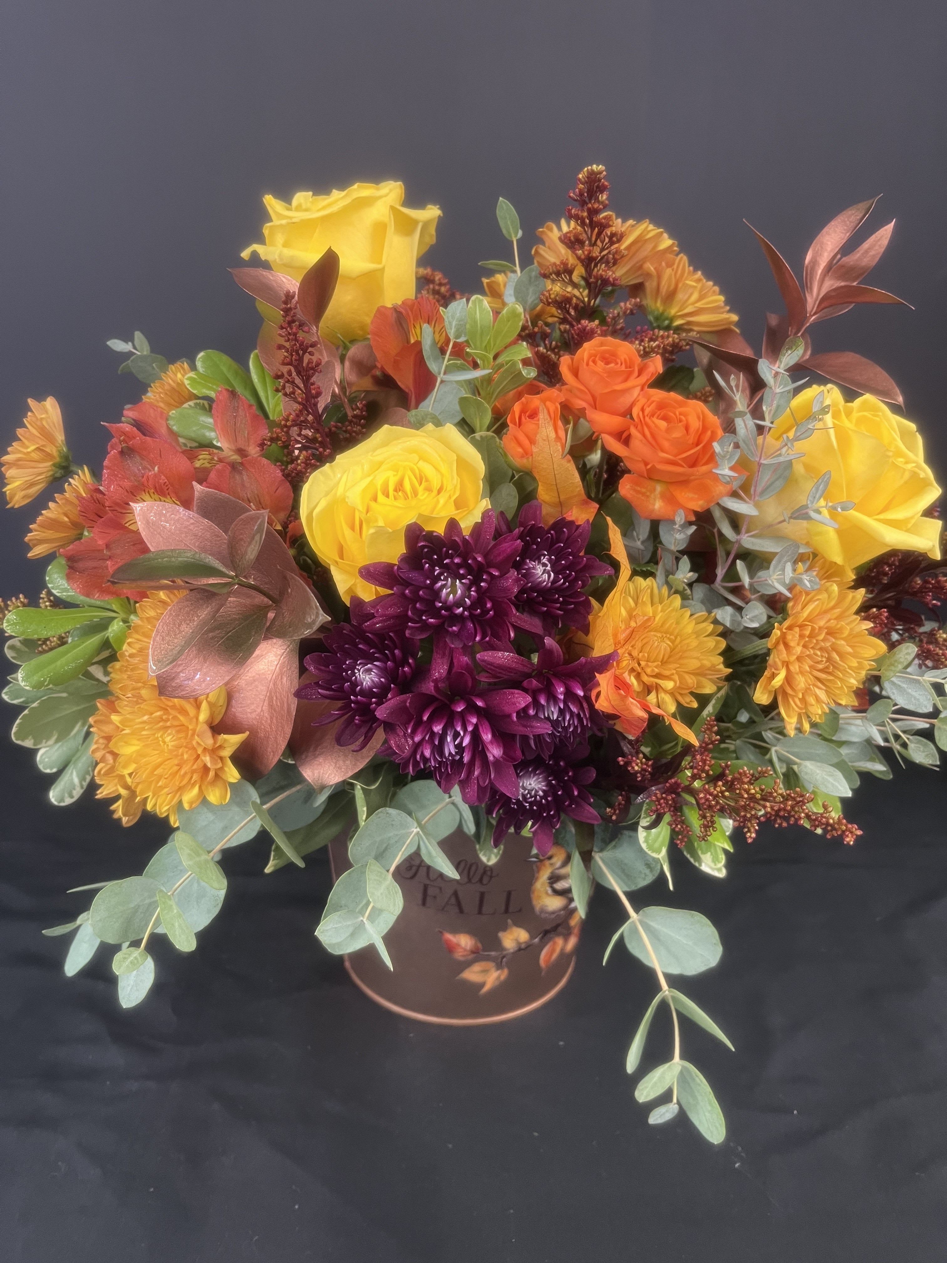 Fall bouquet with yellow roses, orange spray roses, and purple chrysanthemums in a decorative container