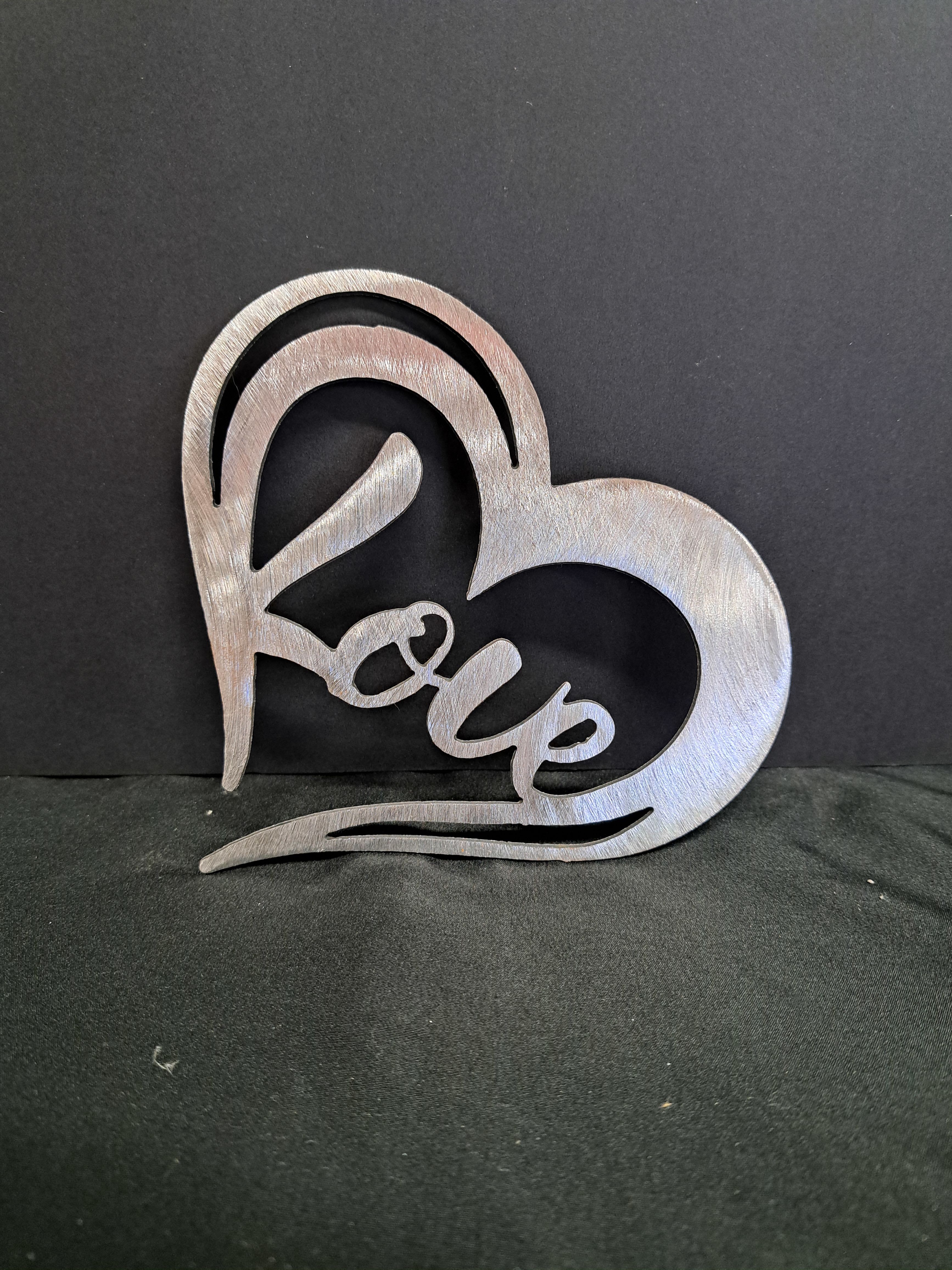 Metal Heart wall hanger Love - A brushed metal heart-shaped wall hanger that spells out "love" brings a modern, romantic accent to any room - perfect as a gift for anniversaries, Valentine's Day, or to mark a special relationship. The reflective silver finish (and an alternative glossy red option) catches light across its flowing cutout lettering and curved silhouette, creating a bold focal point that reads as both ornament and message.  Made from decorative metal with a handcrafted look, this piece is available in striking silver or bright red and is sized to work as a wall or tabletop accent (hangs easily for instant display). The clean lines and durable finish mean it complements contemporary, rustic, or eclectic décor and will make the person you're thinking of feel noticed and cherished.  Choose this heart hanger when you want a lasting, non-floral way to say "I love you" - it's a thoughtful pick for partners, close friends, or to dress up a gallery wall. Care is simple: wipe clean with a soft, dry cloth to preserve the finish and keep the cutout lettering bright.