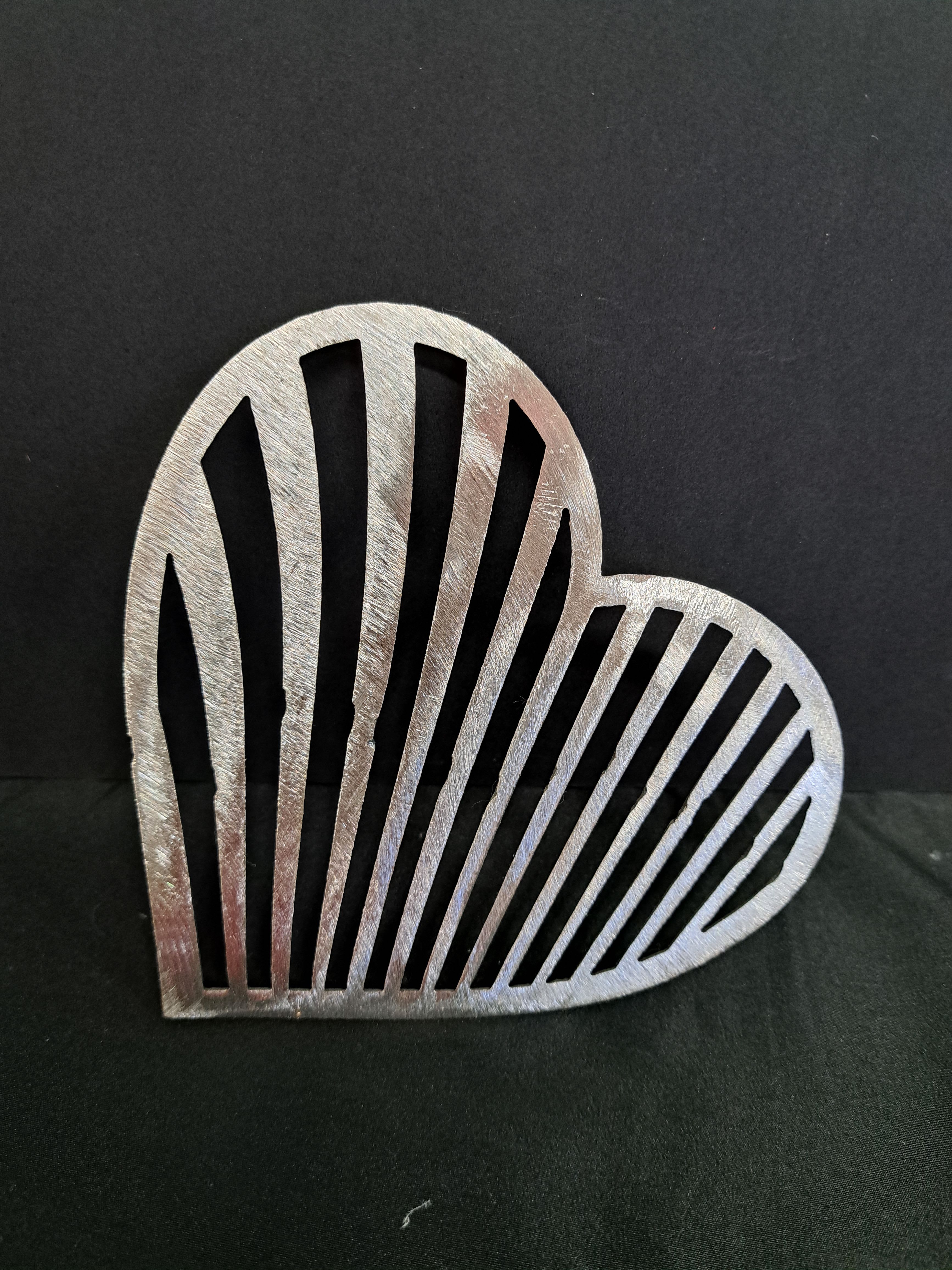 Metal Heart wall hanger - A hand-crafted metal heart wall hanger brings a graphic, romantic touch to any space-perfect as a small anniversary gift or a cheerful Valentine's accent for the person you're thinking of. This decorative heart wall hanger works equally well for everyday home décor or as a simple, heartfelt token to mark birthdays, engagements, or just-because moments.  Choose from a brushed silver-toned finish or a bright orange cutout, each with vertical slotted openings that create a lightweight, openwork silhouette against a wall. The clean lines and subtle texture read modern yet handmade; the piece is metal and designed as a standalone accent rather than a floral arrangement. Sized to fit easily on an entryway, bedroom, or gallery wall, it pairs well with framed photos or other small keepsakes to make the recipient feel seen and celebrated.