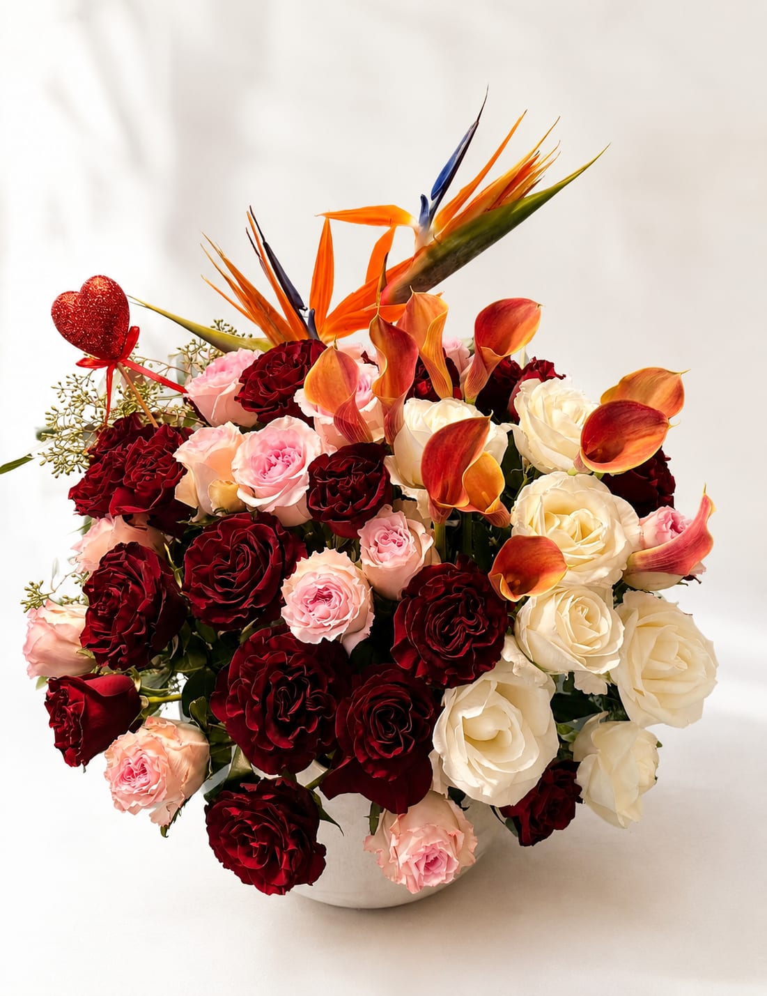 Bouquet of red, pink, and white roses with orange calla lilies in a white vase