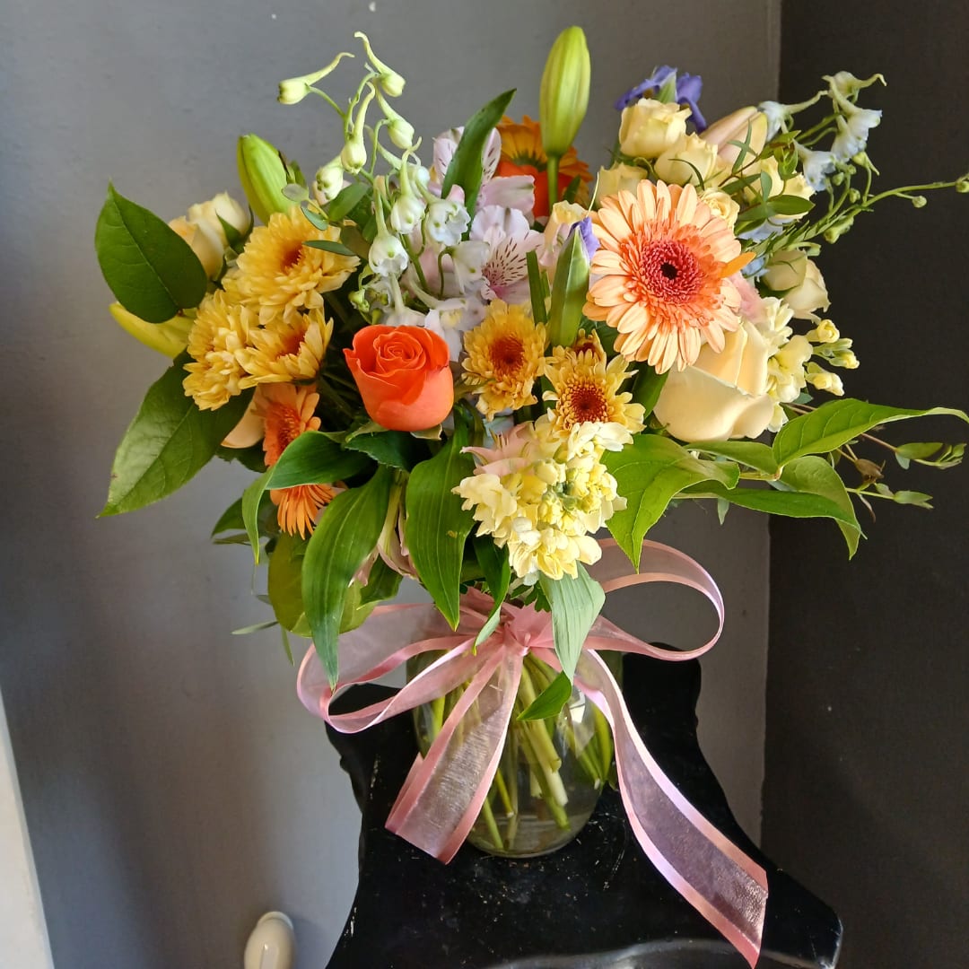 Mixed bouquet of peach, yellow, and white flowers in a glass vase with pink ribbon