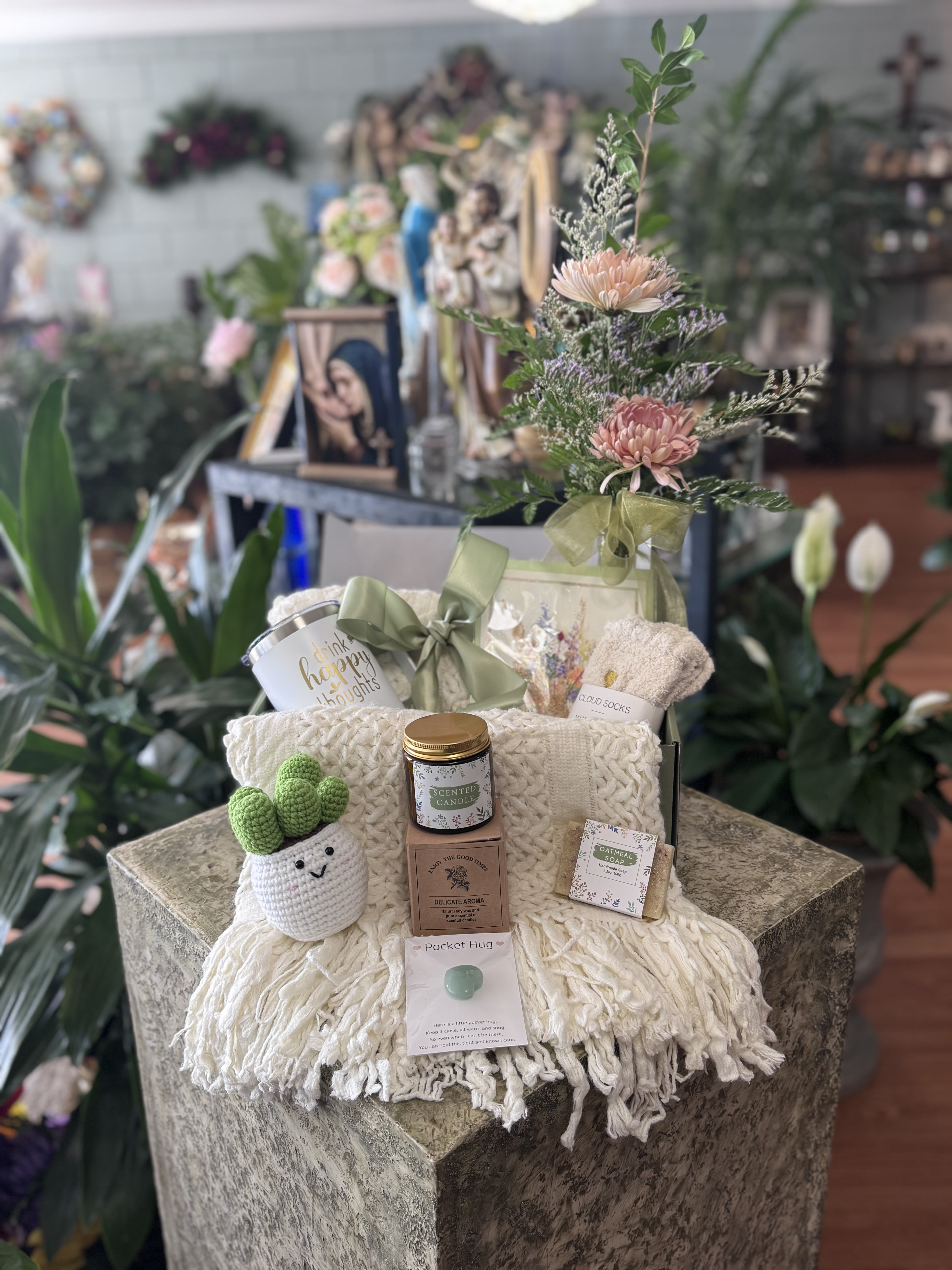 Wrapped In Love - Soft pink chrysanthemums and airy greenery combine with cozy keepsakes to make a gentle Get Well or Thinking of You gift set that feels like a warm hug. This wrapped flower arrangement and gift set is perfect for Mother's Day, Just Because moments, or as a thoughtful get-well surprise. It pairs a small upright bouquet tied with a muted green ribbon alongside a curated basket of comfort items to soothe and uplift the person you're thinking of.  The bouquet features soft pink chrysanthemum blooms, delicate filler stems, and fresh green foliage arranged upright for a tidy, comforting silhouette. The gift basket is layered with a cream knit throw blanket, a scented candle, a soap bar, a boxed pair of socks, a ceramic mug, a crocheted cactus plush, and a thoughtful gift box and ribbon. Everything presented together for an immediate at-home pick-me-up.