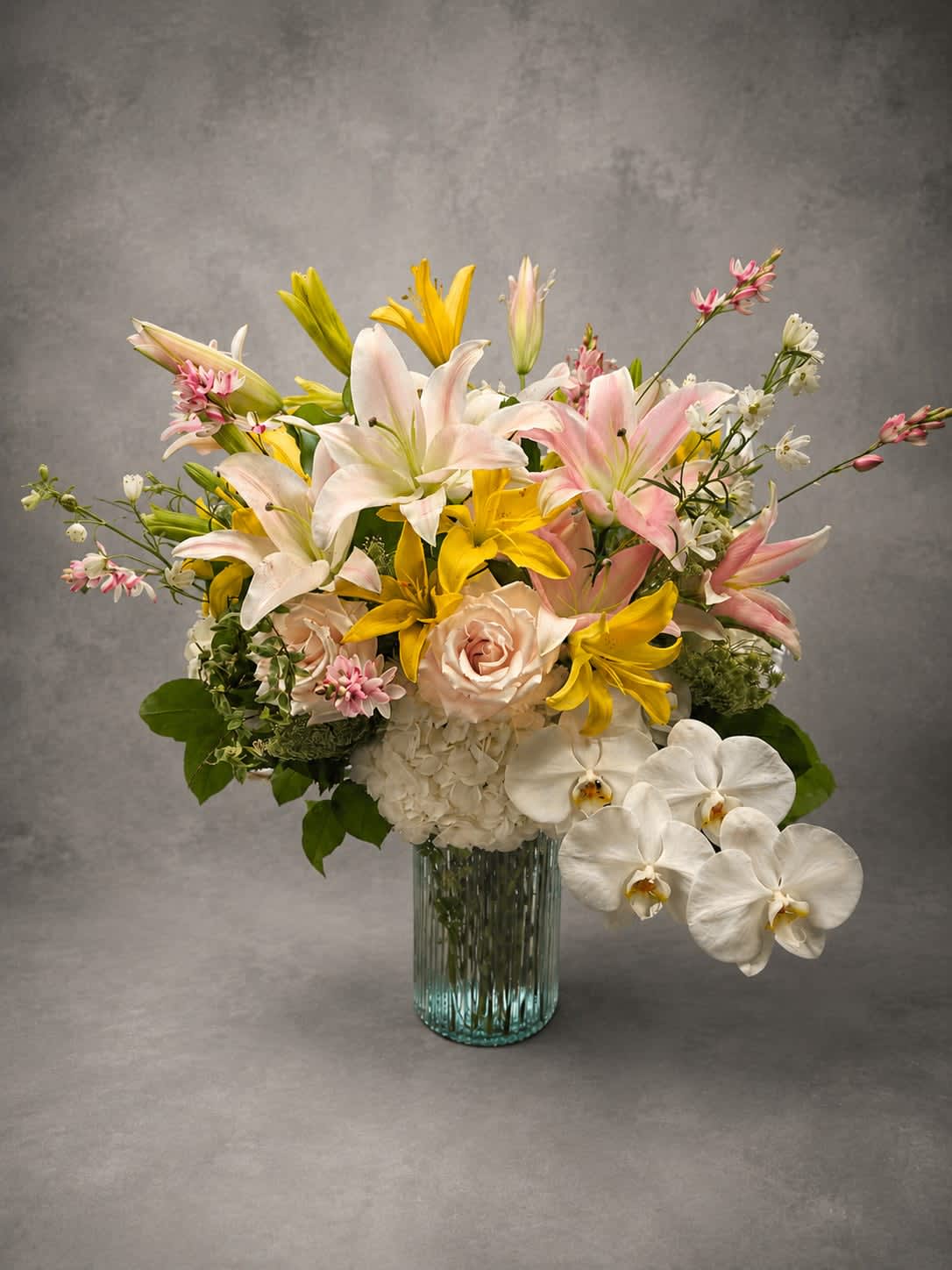 Blushing Lilies - An elegant gathering of premium lilies, blush roses, white hydrangea, and seasonal accents arranged in a modern glass vase. Soft colors and lush textures create a design that feels romantic, refined, and effortlessly beautiful.