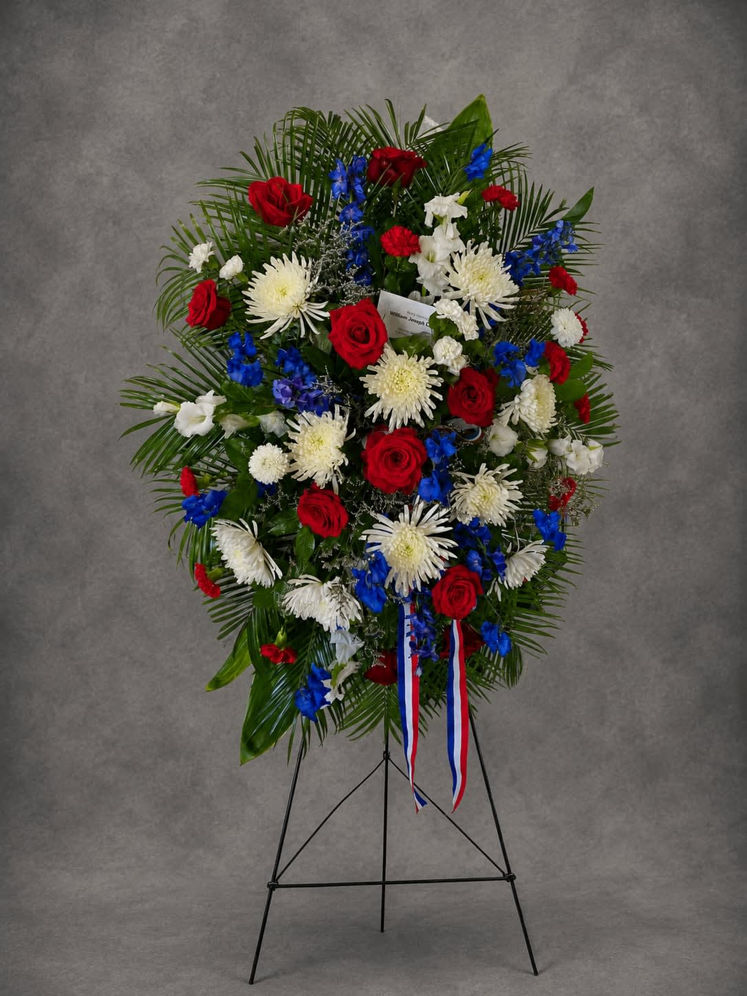 Honor and Valor - A distinguished standing tribute featuring red roses, white chrysanthemums, vibrant blue delphinium, and lush greenery. Designed to honor a life of service, courage, and unwavering dedication, this arrangement offers a heartfelt expression of remembrance and respect.