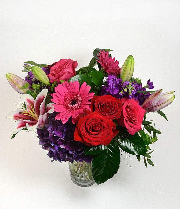 Mixed bouquet of roses, lilies, gerbera daisies, and purple hydrangea in a glass vase