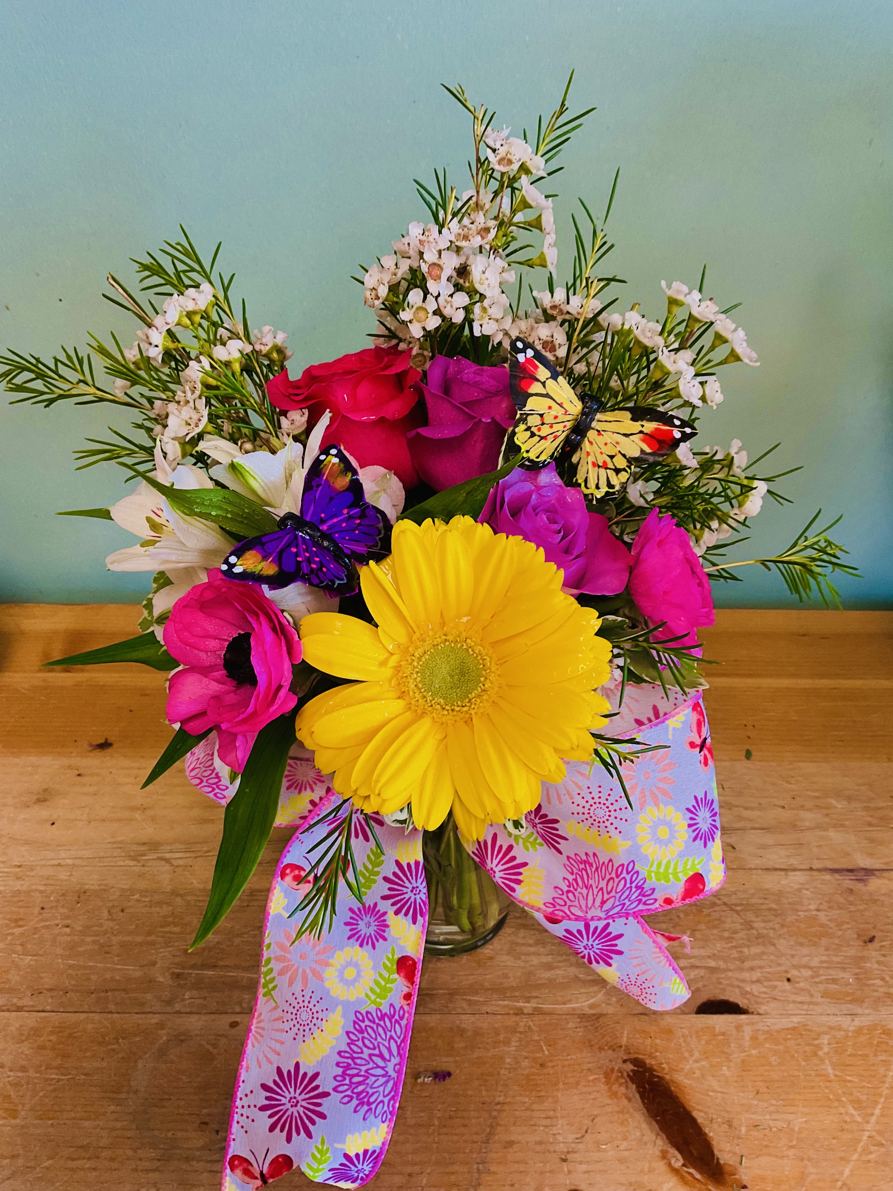 Summer Fun  - A single sun-bright yellow or any color daisy greets the eye in this Summer Fun flower arrangement, a lively bouquet perfect for a birthday, thank-you, or just-because gift. The cheerful mix of pink and magenta roses with crisp white lily-like blooms and purple accents creates an uplifting, celebratory look that will make the person you're thinking of feel noticed and genuinely celebrated.  This glass vase design combines texture and airiness - small white filler flowers and loose greenery give the arrangement a carefree, garden-fresh silhouette while two playful butterfly picks add a whimsical touch. The bouquet is finished with a wide pastel ribbon printed with floral patterns for a gift-ready presentation; tones include bright yellow, soft pinks, white, and touches of purple for contrast.  Send this when you want to brighten someone's day or mark a happy occasion with color and movement. Arrives arranged in a clear glass vase, ready to display; for best life, refresh the water every few days and remove any wilted foliage so the recipient enjoys the arrangement longer.