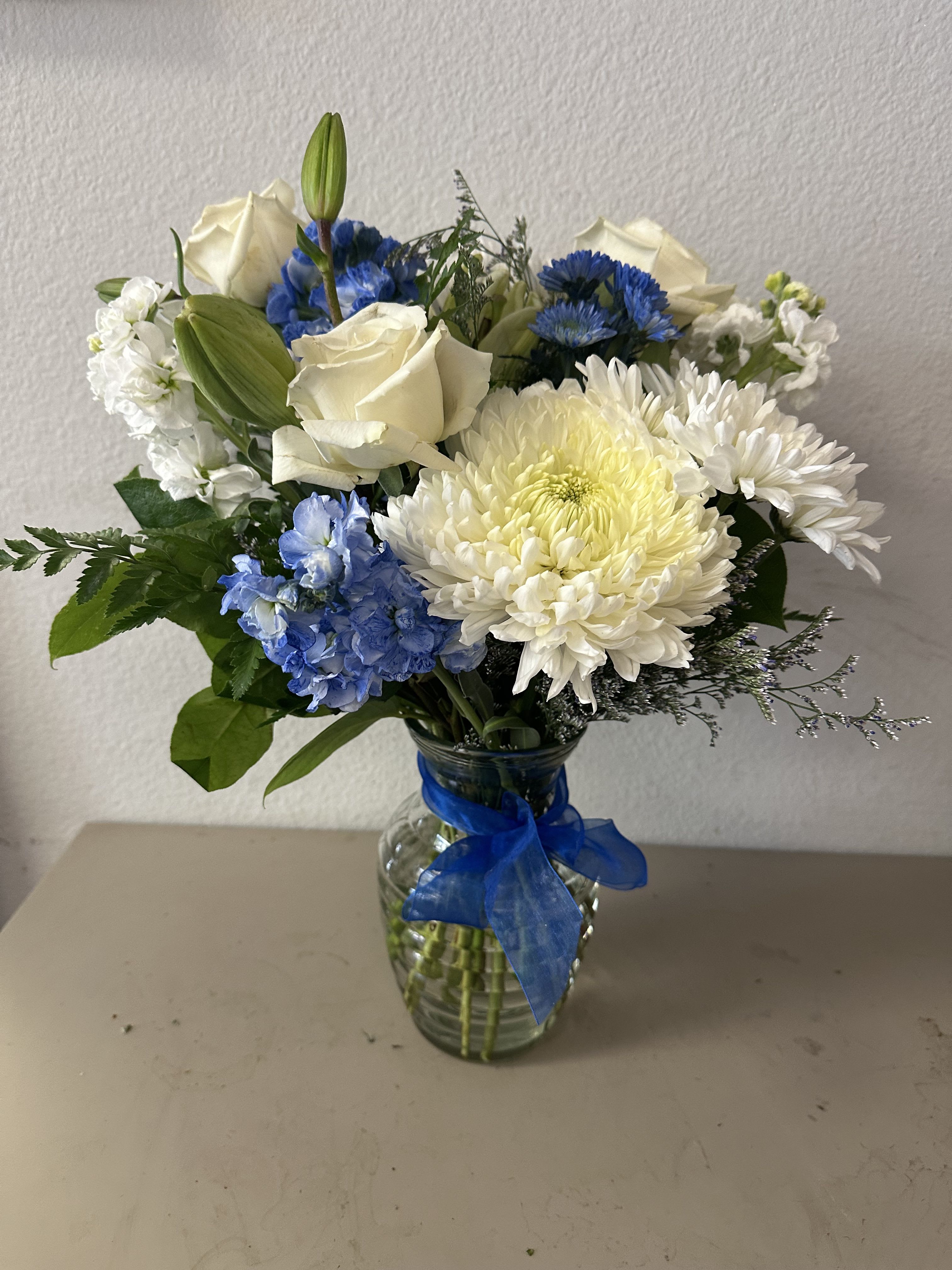 White roses and blue flowers arranged in a glass vase with a blue ribbon.