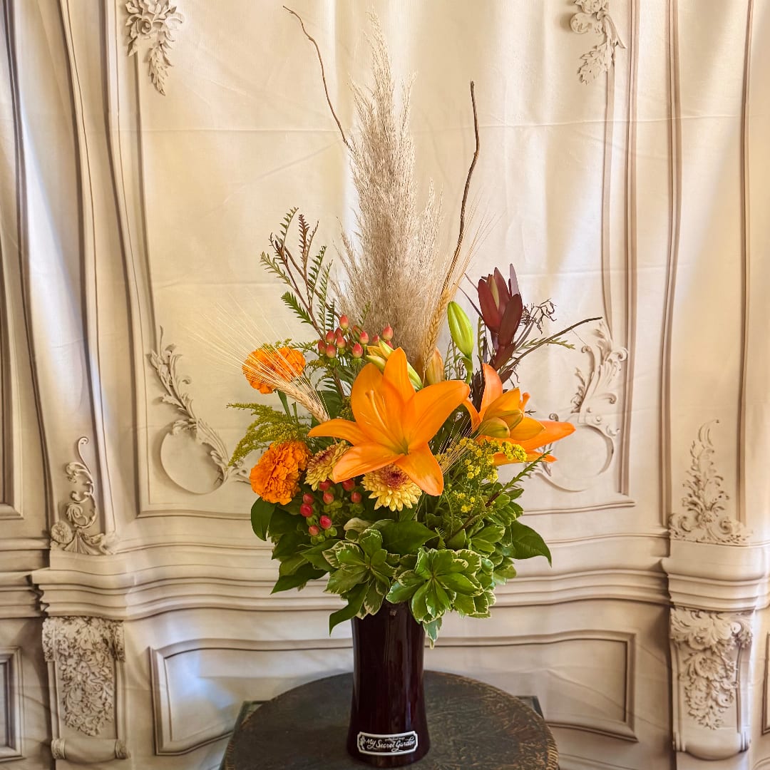 Orange lily arrangement in a tall dark vase with dried grasses