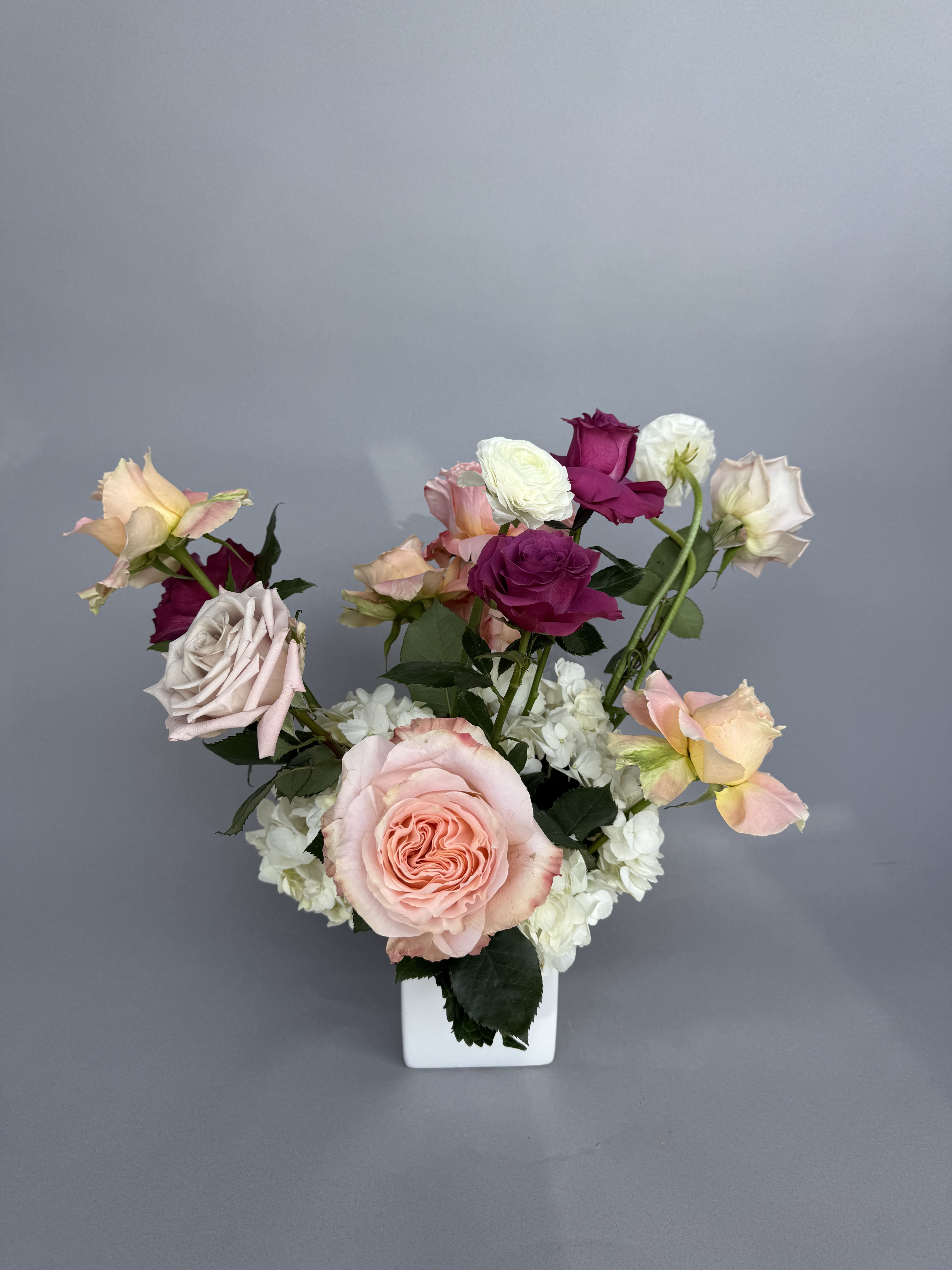 Summer Designer's Choice - A loose, garden-style arrangement that reads like a handpicked bouquet - this Summer Designer's Choice flower arrangement blends hydrangeas, roses, and ranunculus in pink, purple, white, and peach for a soft yet dramatic look perfect for an anniversary or a "just because" surprise. The tall, airy composition sends stems and blooms in varied directions, giving the recipient the feeling of flowers gathered straight from a summer patch: romantic, relaxed, and unmistakably thoughtful.  Clusters of white hydrangea form a cloud-like base, topped with blush pink, peach, ivory and deep magenta roses and a few ruffled white ranunculus for layered texture and depth. Arranged in a clean white ceramic vase, the palette balances pastel softness with saturated color accents so it looks equally at home on a bedside table, dining room, or office desk. Ideal for saying "thank you," celebrating love and romance, or brightening Mother's Day, this arrangement arrives ready to display; keep stems in fresh water, out of direct sun, and enjoy blooms for several days longer with regular trimming.