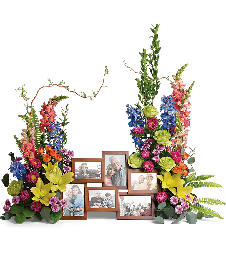 Loving Farewell Photo Tribute - An awe-inspiring accent for your beloved photos at the service, this lush rose, lily and delphinium bouquet reflects a colorful life full of radiant memories. Green roses, orange spray roses, yellow asiatic lilies, blue delphinium, orange snapdragons, hot pink matsumoto asters, and lavender button spray chrysanthemums are accented with curly willow, myrtle, sword fern, silver dollar eucalyptus, and lemon leaf. Arrangement does not include picture frame. Approximately 40" W x 33 1/2" H 