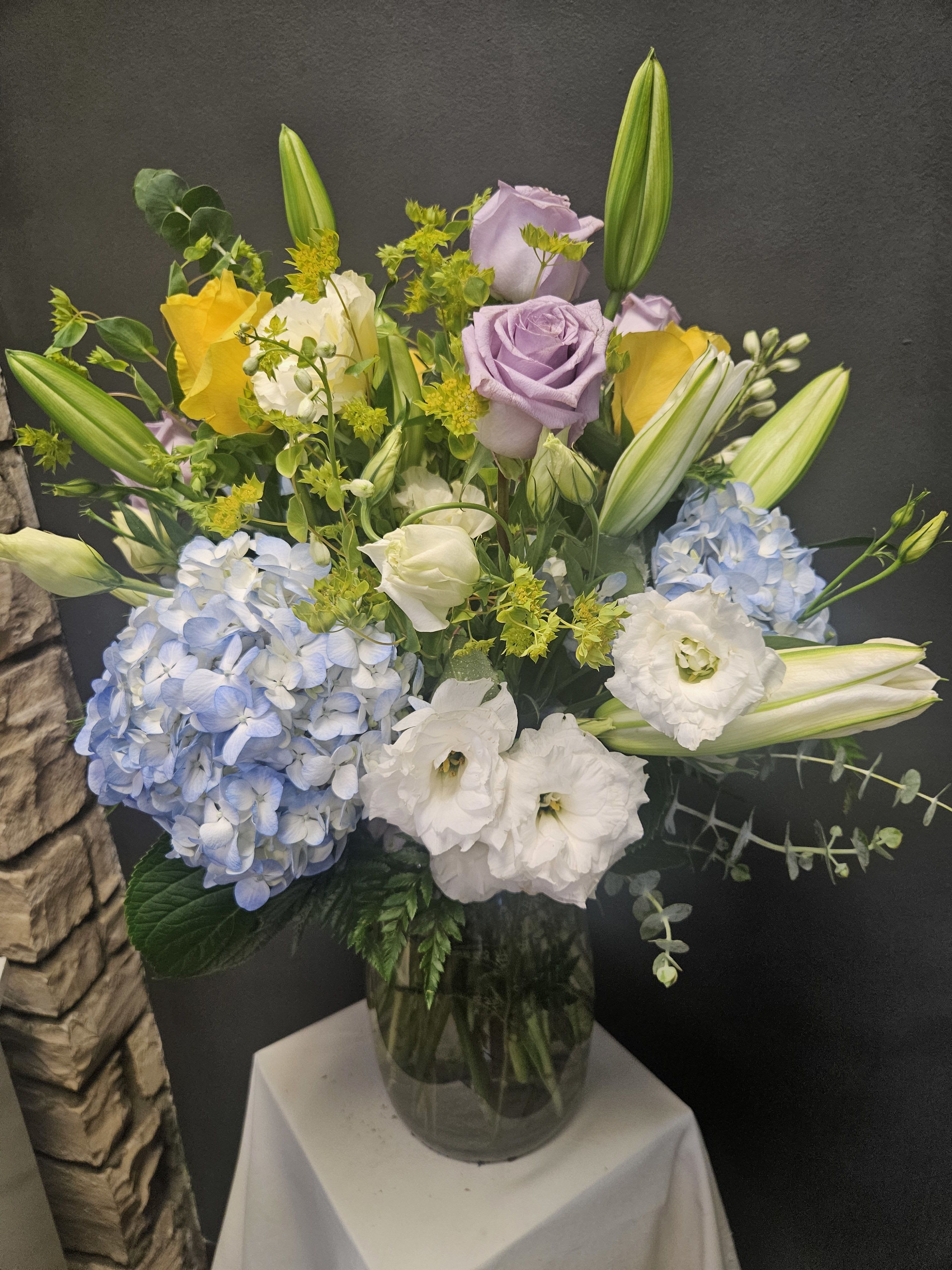 Mixed bouquet with blue hydrangeas, roses, lilies, and white blooms in a glass vase