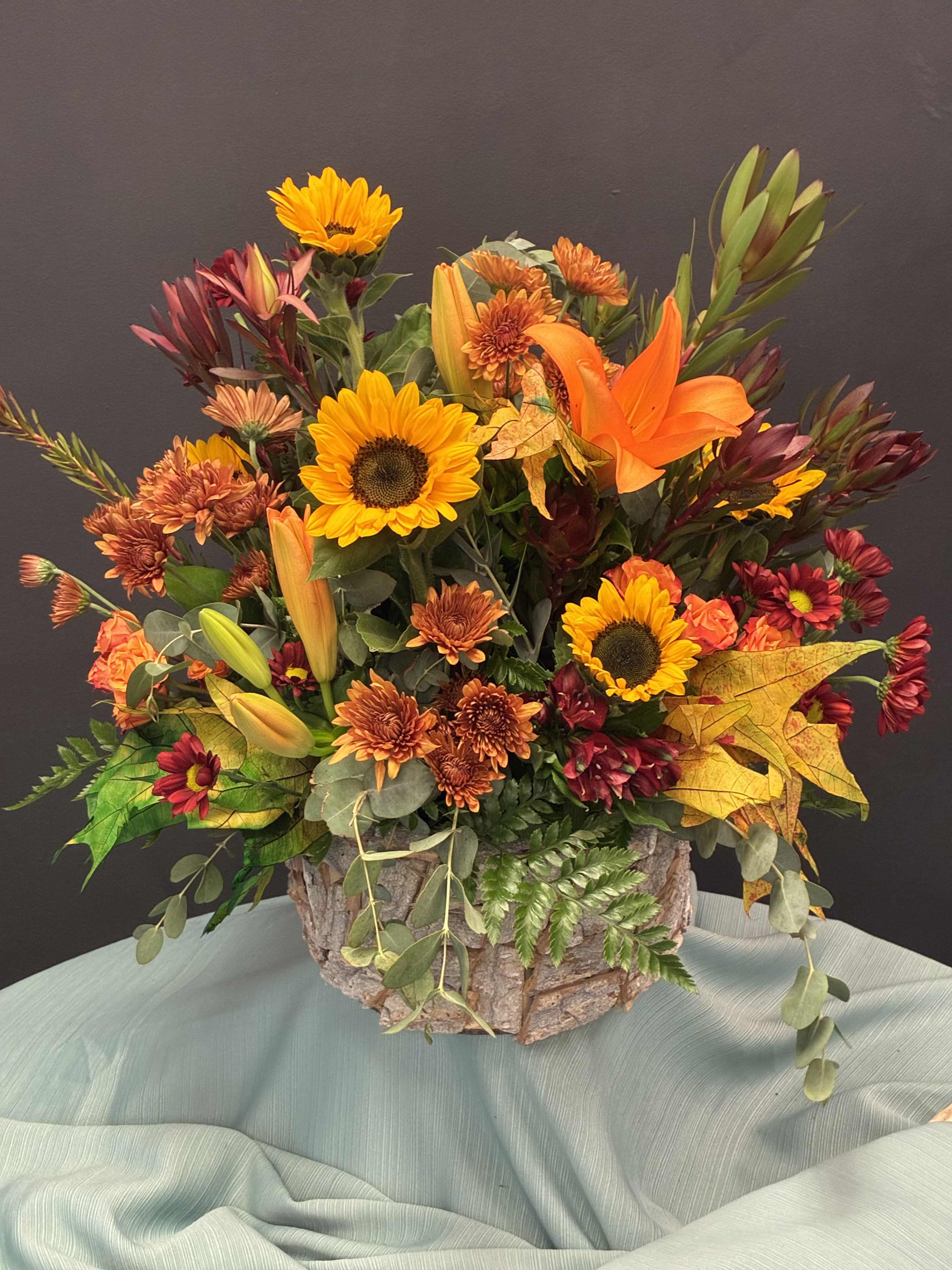 Maple Glow - A graceful ode to autumn’s beauty. Sunflowers and lilies bloom in harmony, cradled by rustic bark and kissed by golden leaves. This arrangement whispers warmth, gratitude, and the quiet magic of the season.