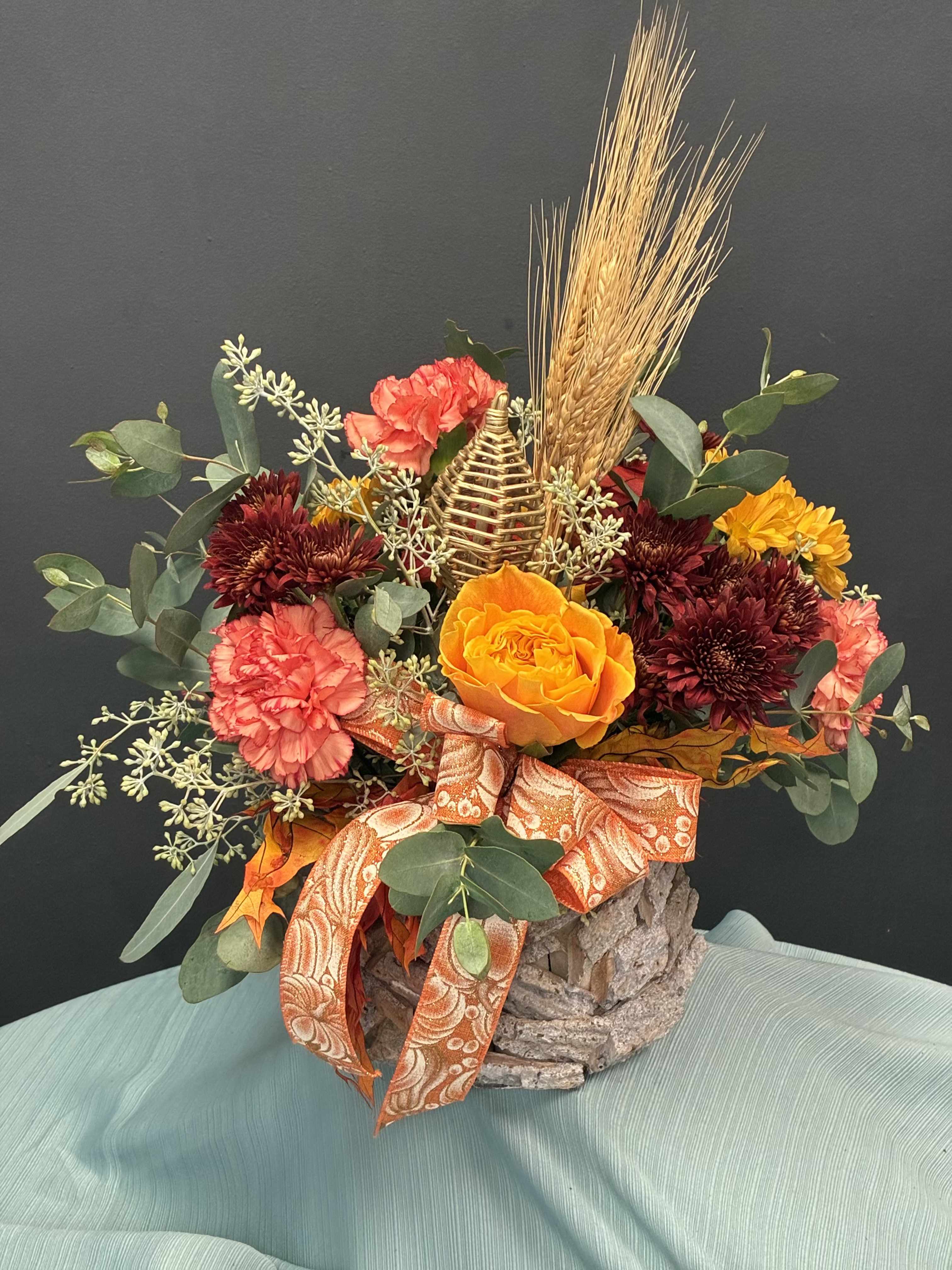Amber Nest  - A rustic embrace of autumn’s gentler side. Marmalade roses and coral carnations bloom with quiet warmth, while maroon chrysanthemums and golden wheat add depth and texture. Eucalyptus leaves and baby’s breath soften the palette, and a pine cone nestled among the blooms whispers woodland charm. Tied with a paisley ribbon and cradled in bark, this arrangement feels like a cozy invitation to linger in the season’s glow