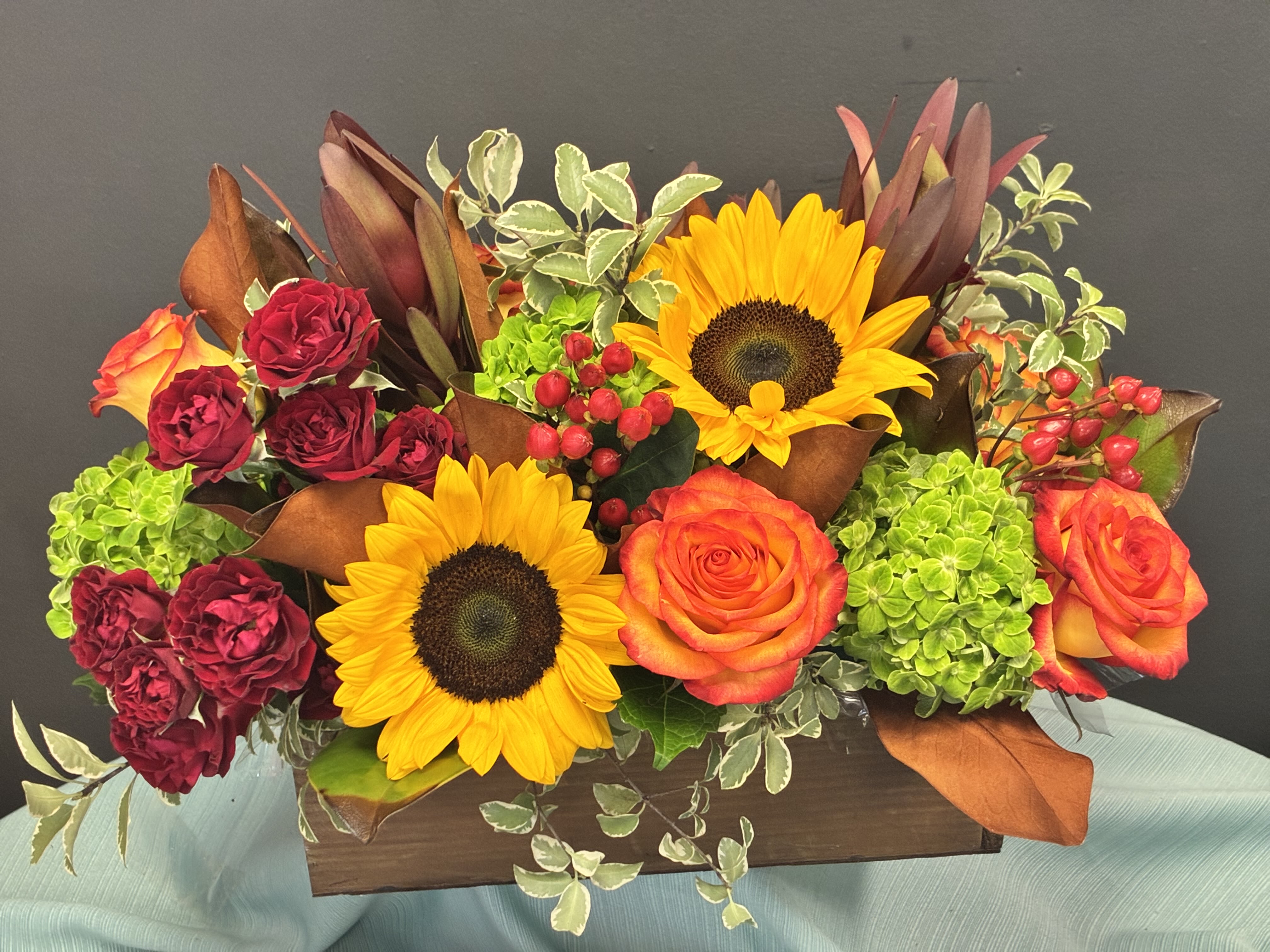 Autumn Sonata - A bold, autumn-inspired centerpiece overflowing with texture and color. Three radiant sunflowers anchor the design with golden warmth, surrounded by fiery orange-red roses and velvety deep red carnations. Clusters of green hydrangea and red hypericum berries add contrast and richness, while variegated greenery and magnolia leaves bring depth. Tall, rust-toned foliage rises above, adding drama and vertical movement. Set in a rustic wooden container, this arrangement feels both festive and grounded—perfect for seasonal gatherings or a warm welcome home.