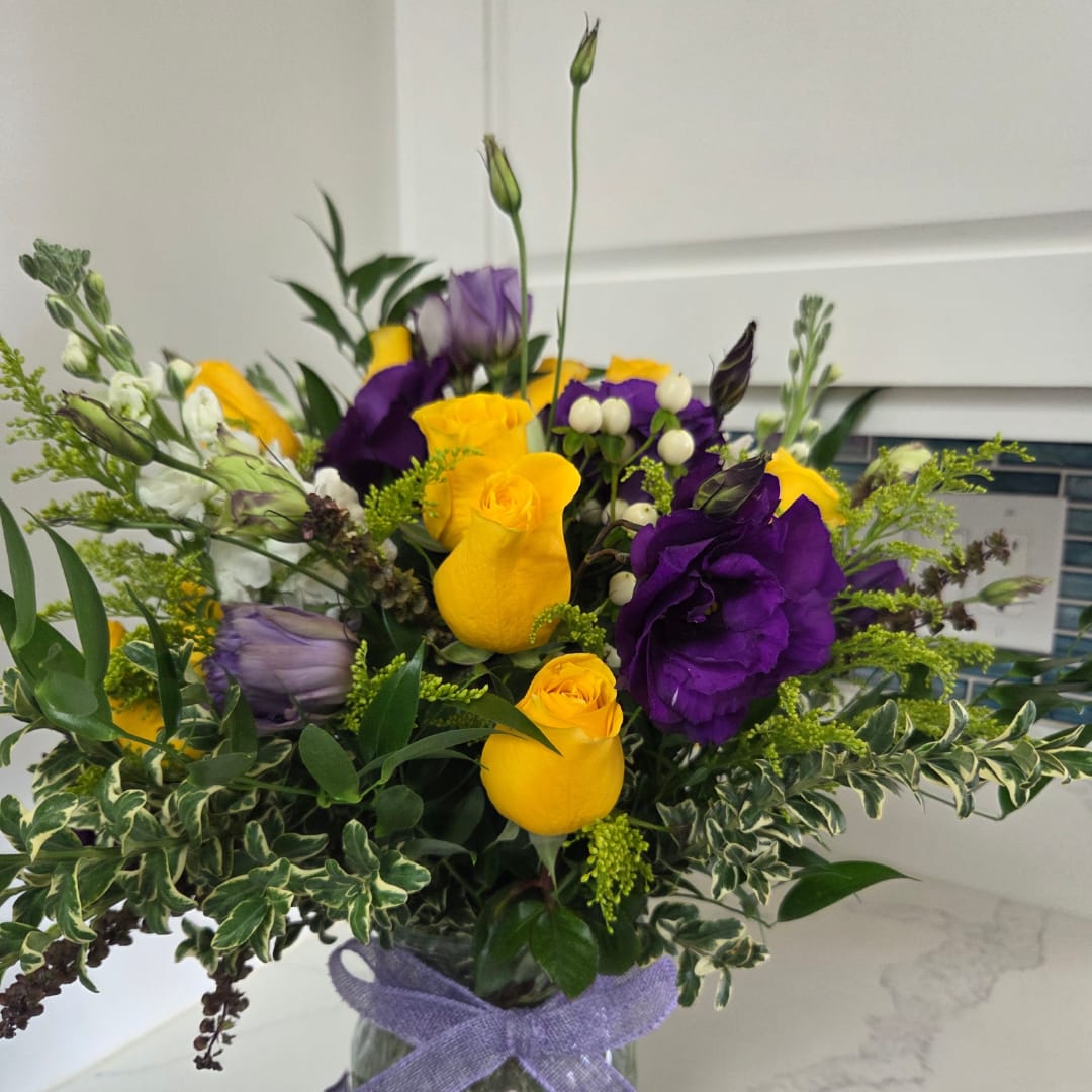 Yellow and purple roses arranged in a glass vase with a lavender ribbon