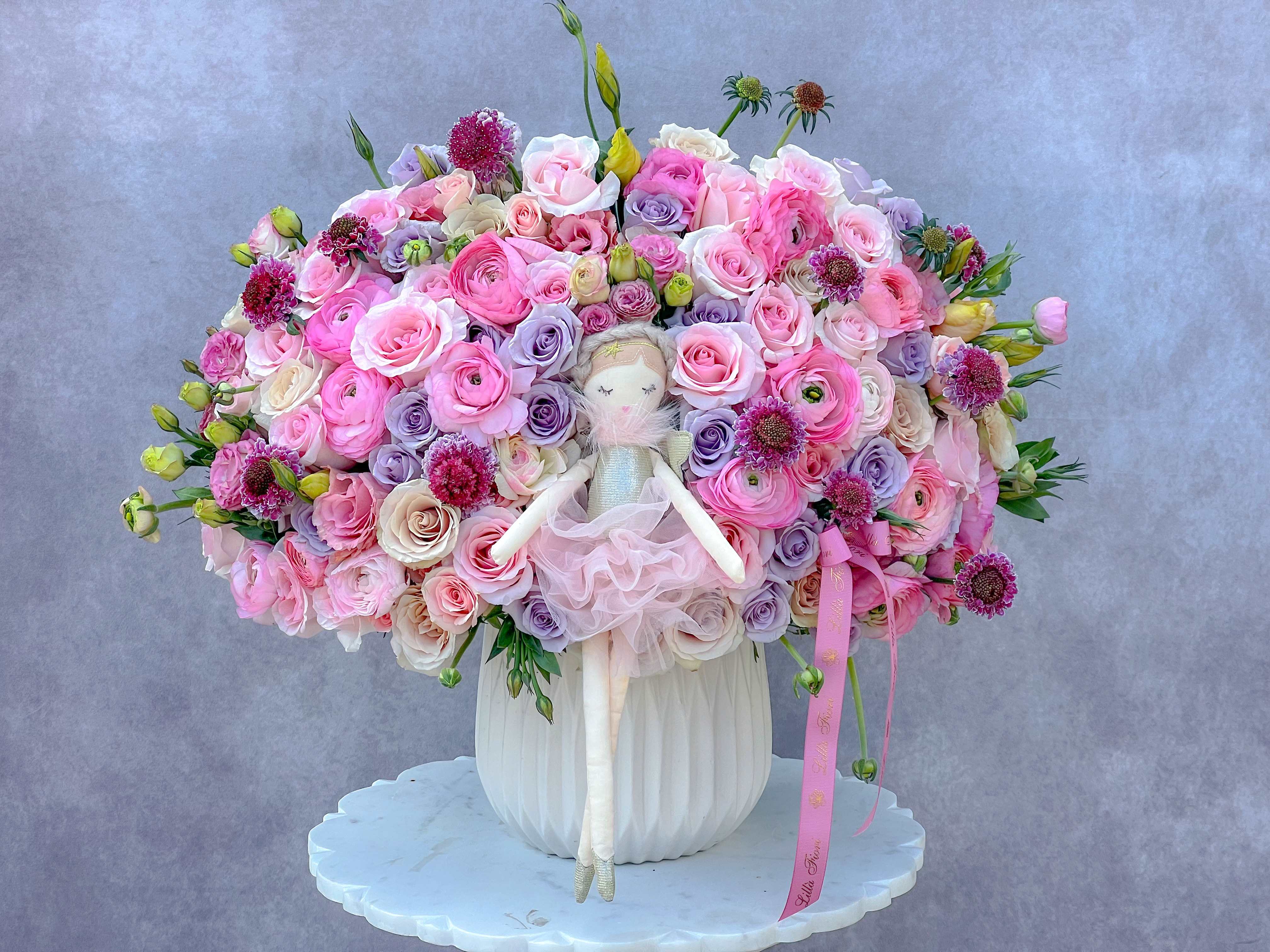 Large pink and lavender rose arrangement with a doll in a white vase