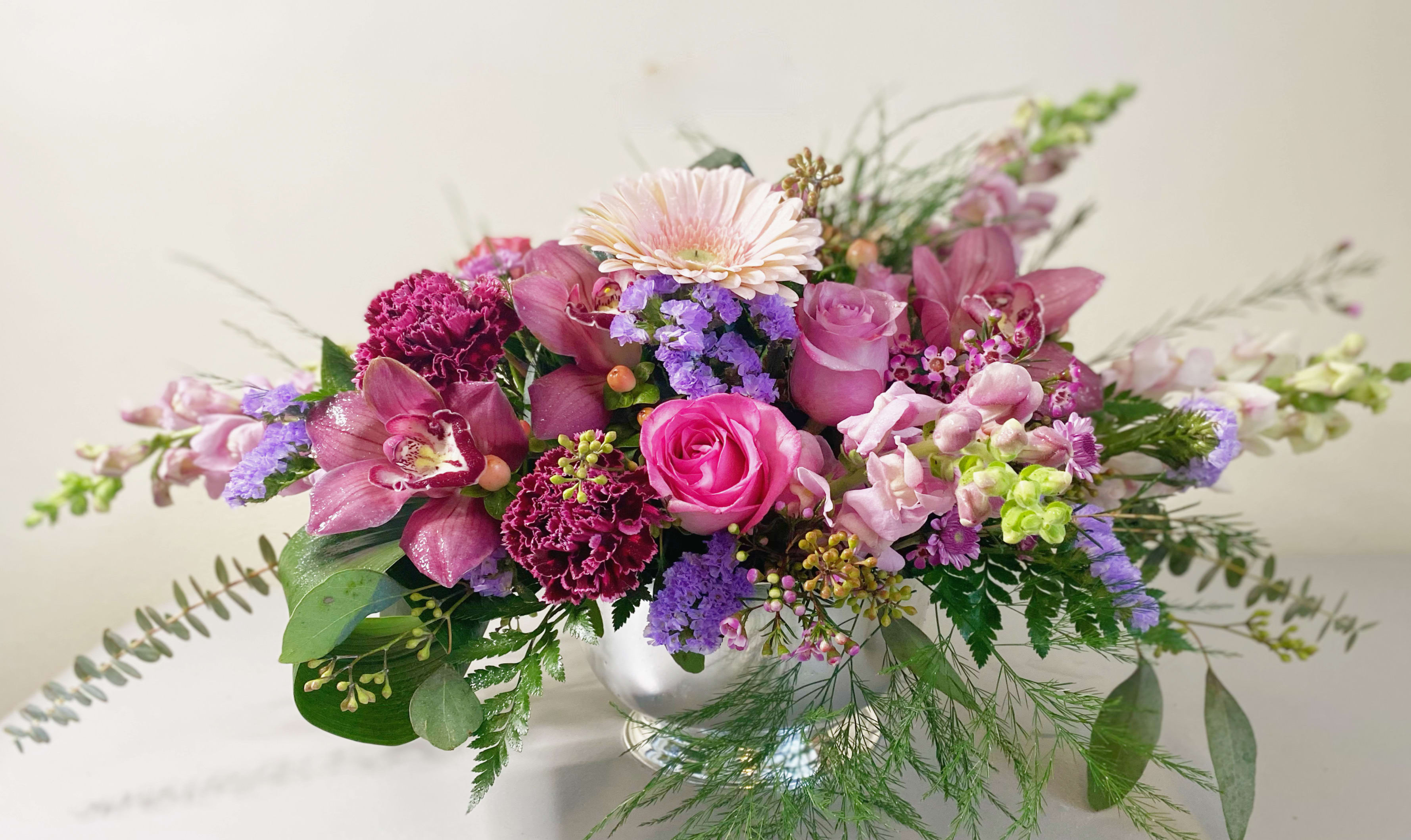 Serene blossoms - Serene blossoms is lush, botanical, soft and beautiful floral arrangement. Pink lavender, burgundy and peach with touch of a garden feel with mixed foilage.Has a stylized and botanical feel with roses, orchids, rolled leaves in a silver container. Approximately 18-20 inches long, 8-10 in wide. 12 to 15 inches high. Perfect for Valentine’s Day or gift for a dinner table. 