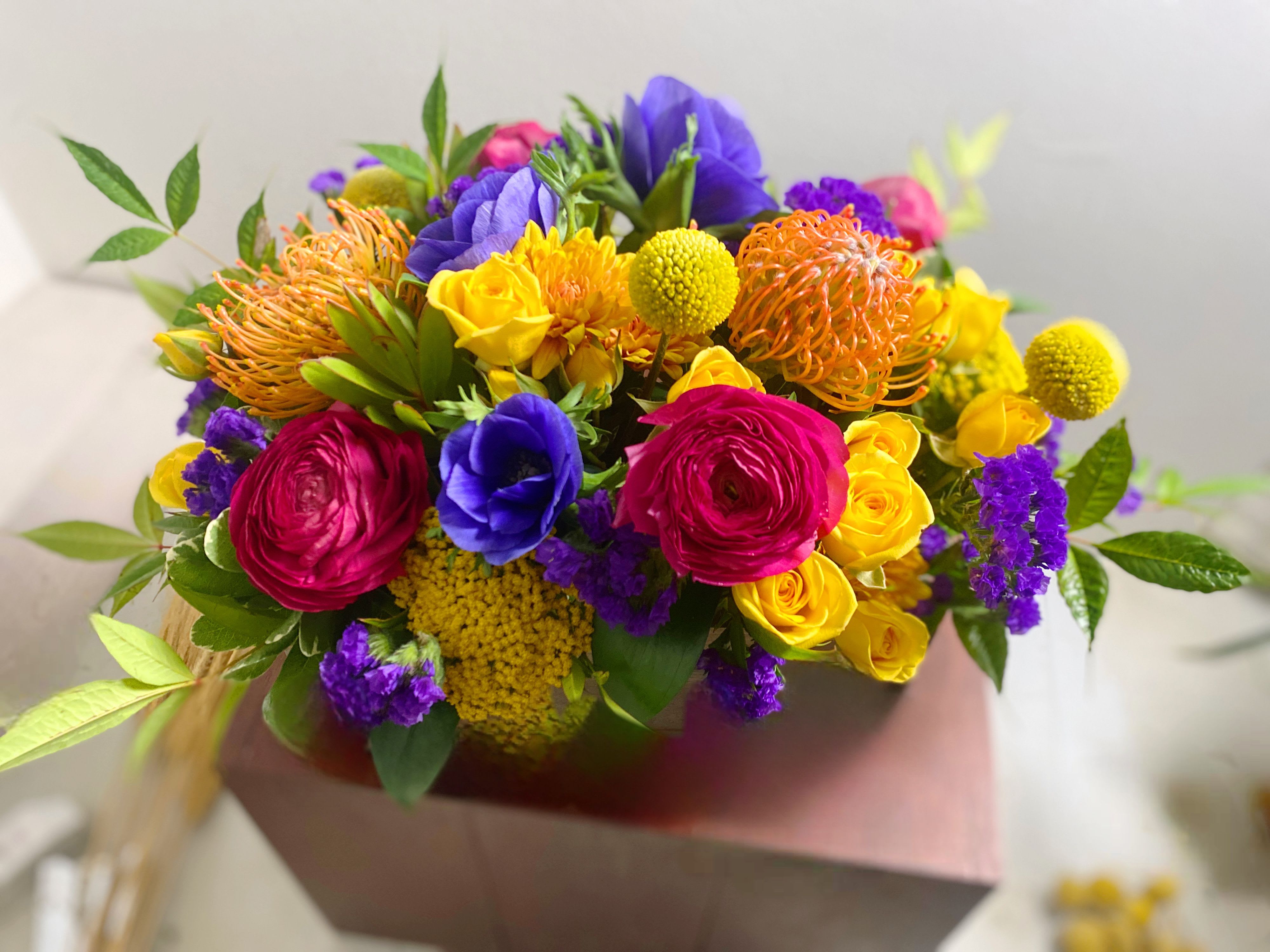 Neon Meadows - Bring a striking architectural edge to your space with the Neon Meadow Bouquet, beautifully arranged in a sleek, low-profile rectangular clear glass vase. Designed exclusively for Indy joy blooms, this unique presentation transforms vibrant, untamed meadow textures into a sophisticated, modern table centerpiece.By utilizing a low rectangular silhouette, our designers can tightly layer the exotic orange pincushion proteas, velvety hot pink ranunculus, and jewel-toned purple anemones side-by-side. Sunny yellow roses and whimsical yellow billy balls sit perfectly at eye level, offering a dense, compact burst of color from every angle.Because it sits low to the surface, this design is the ultimate dinner party conversation piece—stunning your guests with a spectacular view without ever blocking their sightlines.Product Details:Premium Stems: Exotic Orange Pincushion Proteas, Deep Pink Ranunculus, and Electric Purple AnemonesBright Accents: Yellow Cluster Roses, Yellow Craspedia (Billy Balls), and Royal Purple StaticeVessel: High-clarity, low rectangular clear glass vaseDesign Profile: Horizontal pave style, perfect for dining room tables, kitchen islands, mantlepieces, or low coffee tables.Perfect For: Upscale dinner parties, modern housewarmings, stylish thank-you gifts, or high-end office reception desks. 