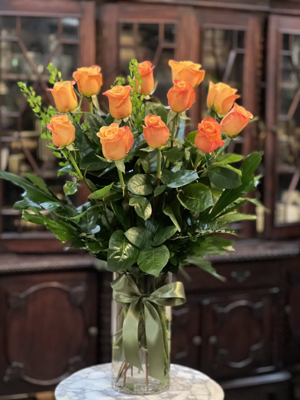 Tall arrangement of orange roses in a clear glass vase with a green ribbon bow