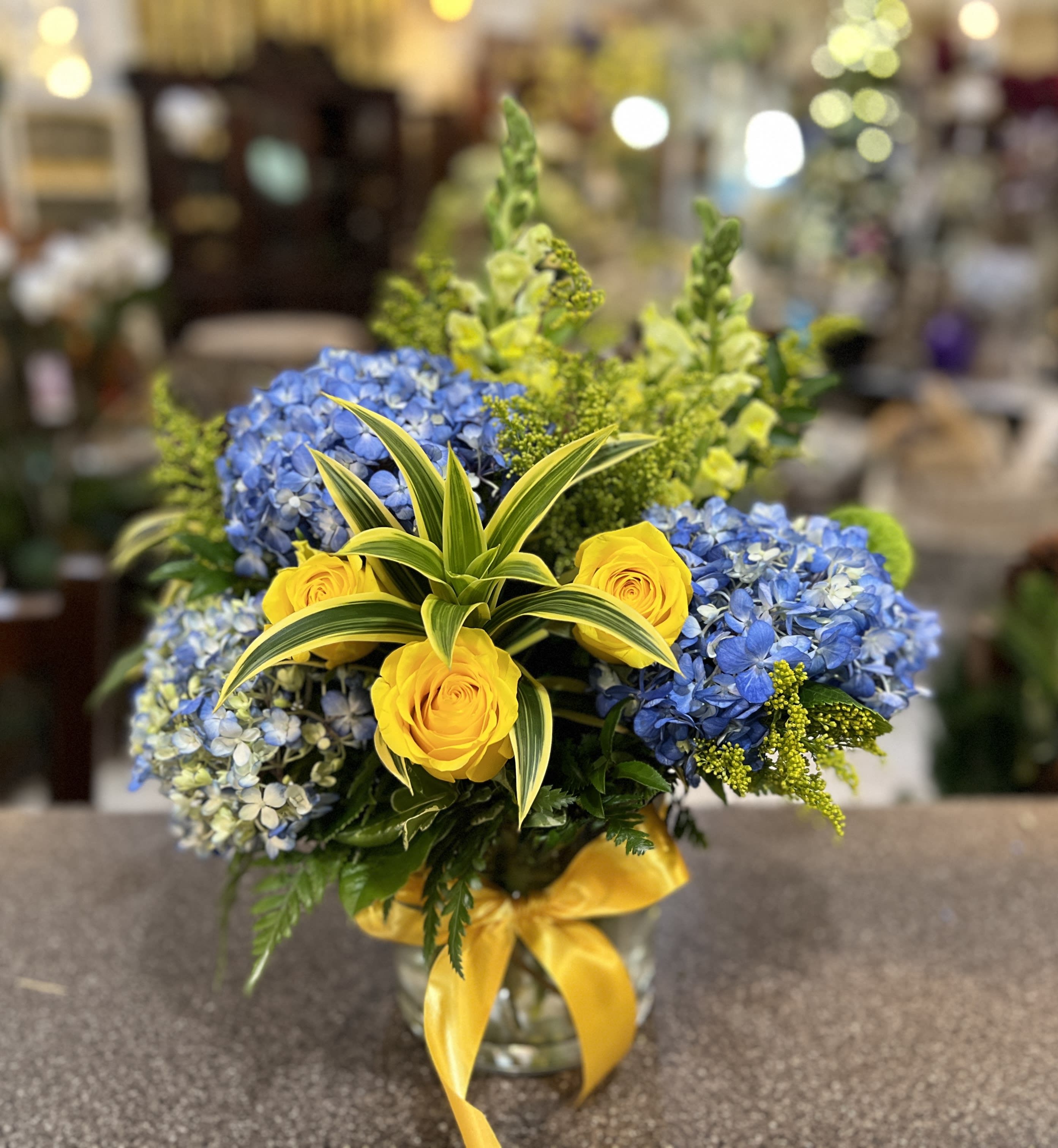 Blue hydrangeas and yellow roses in a glass vase with a yellow ribbon