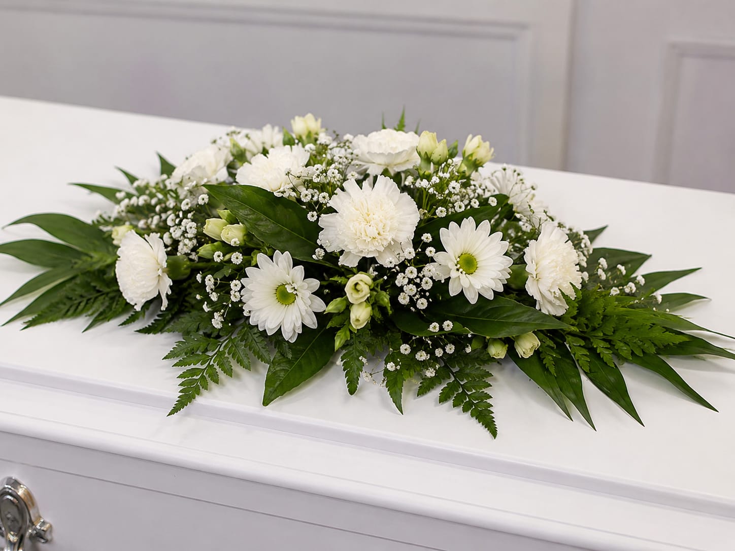 Small Casket Spray - A low, elongated white casket spray arranged to convey quiet reverence-the kind of sympathy arrangement that helps the person you're thinking of feel honored and remembered. This small casket spray is budget friendly, and ideal for funeral and memorial services, resting across the casket with a calm, all-white palette that reads respectful and serene.  The spray is layered over broad, glossy foliage and delicate fern-like greenery for a structured, formal silhouette. Its horizontal, full-coverage shape provides a neat, finished presentation that reads well in photos and beside other floral tributes.  Made to comfort mourners and acknowledge a life with grace, this arrangement is best for close family members or intimate services where a smaller yet complete casket tribute is appropriate. Standard care: keep flowers cool and away from direct heat; if delivery or placement details are needed for the service, please let us know when you order.