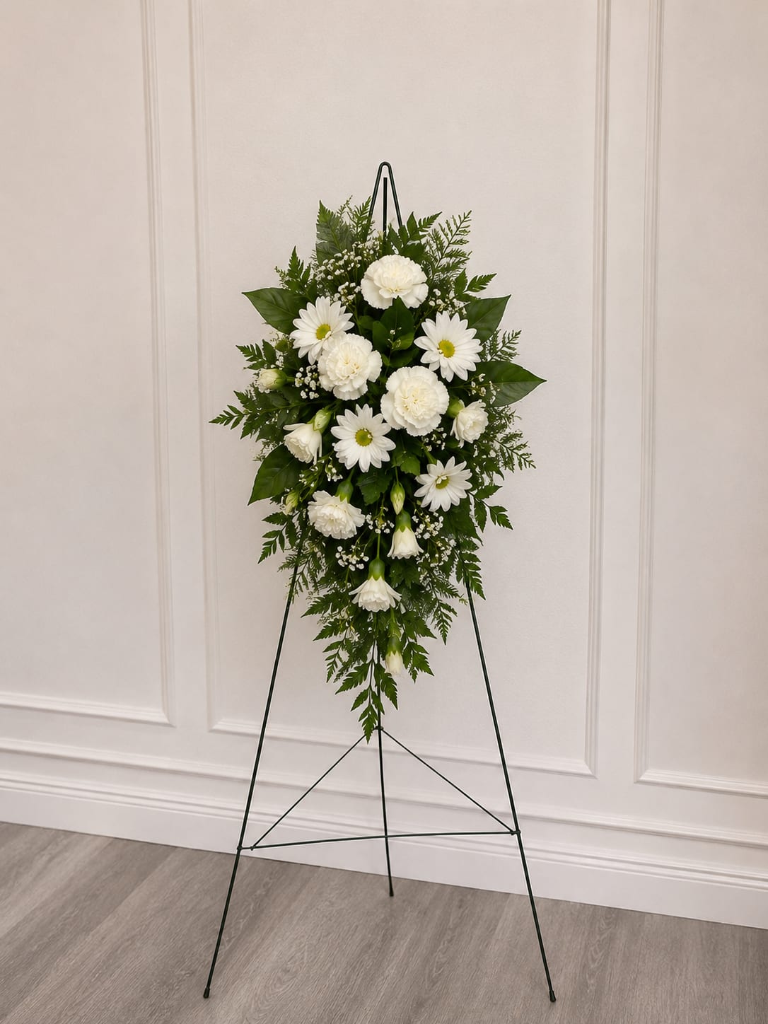 Small White Hanging Spray - All-white standing spray arranged on a metal easel creates a calm, respectful tribute perfect for a sympathy or funeral arrangement. The Small White Hanging Spray reads as a dignified expression of condolence-the person you're remembering will be honored by the serene white  fillers arranged in a tapered, standing design.  Dense ferns and leafy greenery frame the bouquet, giving the spray a full, textured silhouette that carries well when displayed at a service or graveside. The neutral white and green palette keeps the focus on quiet beauty and remembrance, while the black metal easel offers sturdy support and a classic presentation.  Choose this budget friendly spray to convey sympathy, send condolences after a loss, or place at a memorial service-it's sized for chapel aisles and graveside displays. No special care is needed beyond standard floral upkeep; if arranging delivery, confirm service times so the spray arrives for the ceremony.  Stand is 60"; spray size varies depending on flowers used- roughly 1.5' x 2.5'