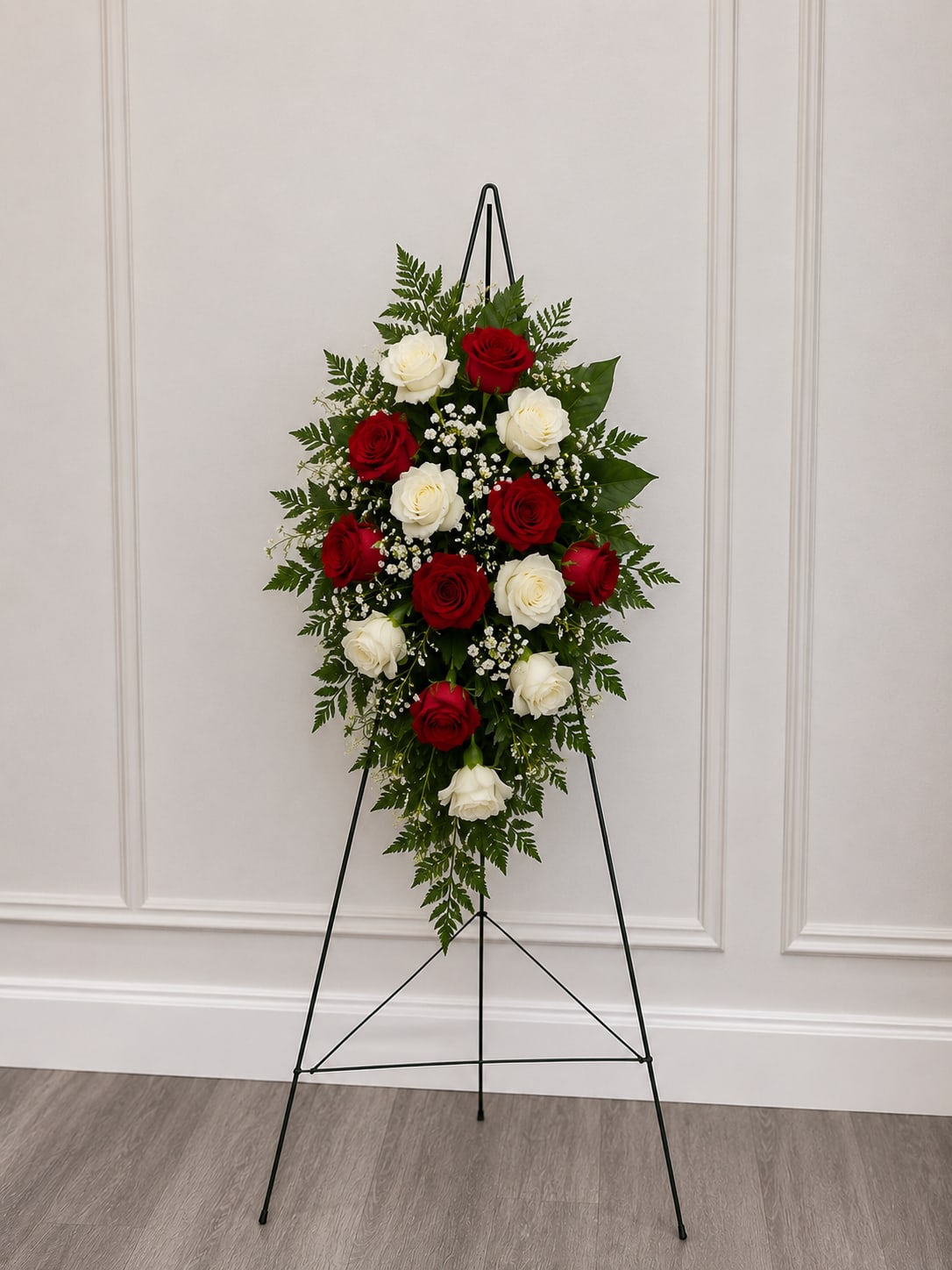 Small Rose Hanging Spray - Alternating bands of red and white roses create a dignified, classic sympathy budget friendly arrangement that reads clearly from a distance - perfect as a funeral standing spray or memorial tribute. The elongated, symmetrical design says "respect and remembrance" in a tasteful way, making the person you're honoring feel seen and remembered.  Tightly clustered red and white roses run down the center, accented with delicate white filler blooms and fern-like greenery that taper to a gentle point at the base. The palette of deep crimson, pure white, and lush green keeps the look formal and reassuring. The arrangement is presented on a sturdy metal easel for easy display beside a casket or at a memorial service.  For sending condolences or marking a memorial, choose this smaller standing spray when you want a meaningful, understated tribute. Standard care is minimal since it's intended for short-term display; the easel is included for convenient placement at the service.  Stand is 60"; spray size varies depending on flowers used- roughly 1.5' x 2.5'