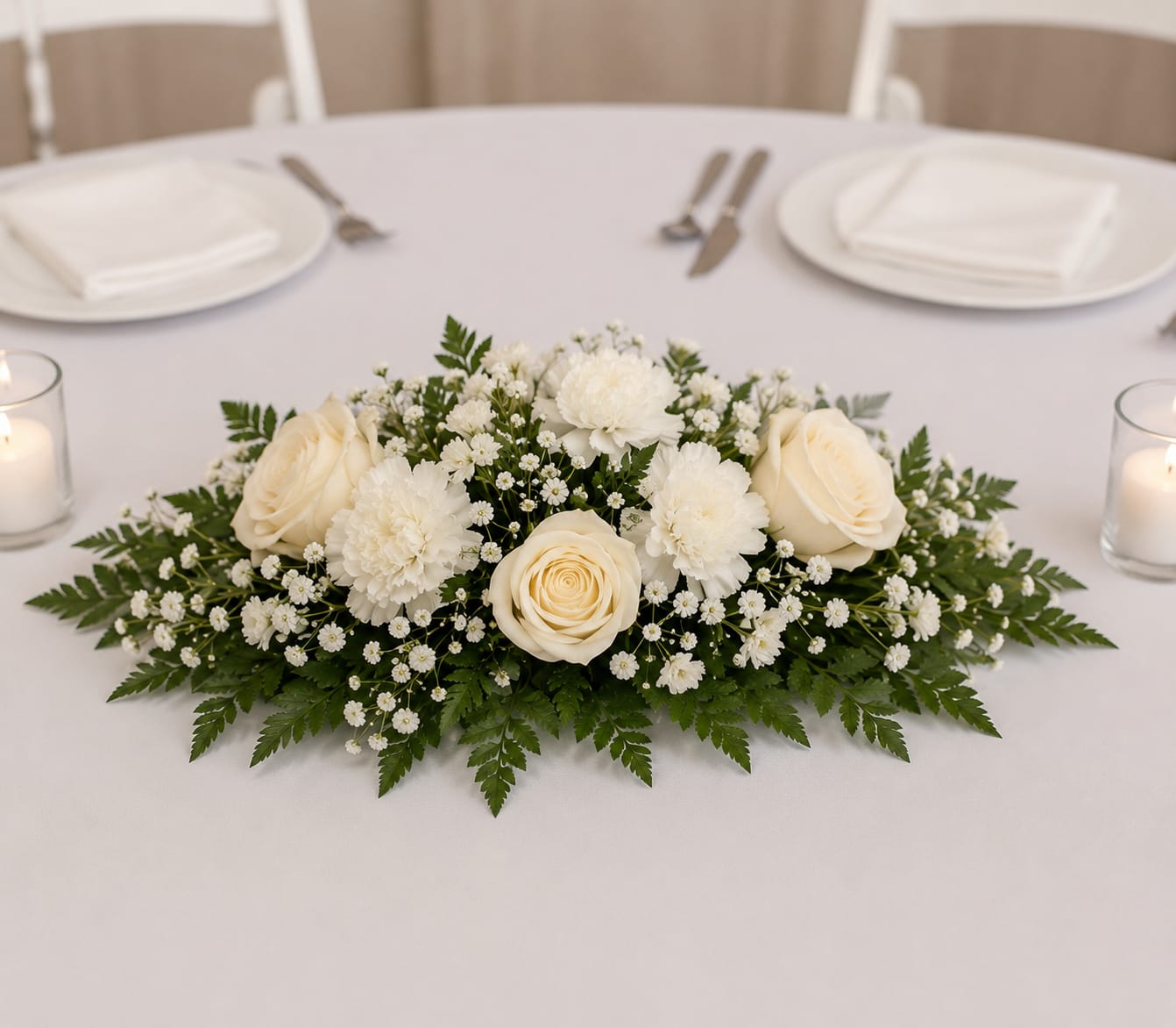Small Table Centerpiece - White flowers arranged low and elongated create a calm, elegant wedding centerpiece that reads formal and romantic-perfect as a small table centerpiece for a wedding reception or other refined event. The gentle palette of white and green keeps the focus on the recipient's moment: she'll feel seen and celebrated by a centerpiece that invites quiet conversation and candlelight.  This budget friendly design uses white floral blooms framed by dense, fern-like greenery that spreads outward across the table for a balanced, full silhouette. The arrangement sits low so guests can see one another while the blooms set a soft, luminous mood.  Ideal for head tables, guest tables, or intimate anniversary dinners, this small centerpiece brings polished charm without overpowering the setting. Because it's compact and low, it works well in tight tabletops and is easy to pair with your chosen place settings; refresh water as needed to keep blooms at their best.