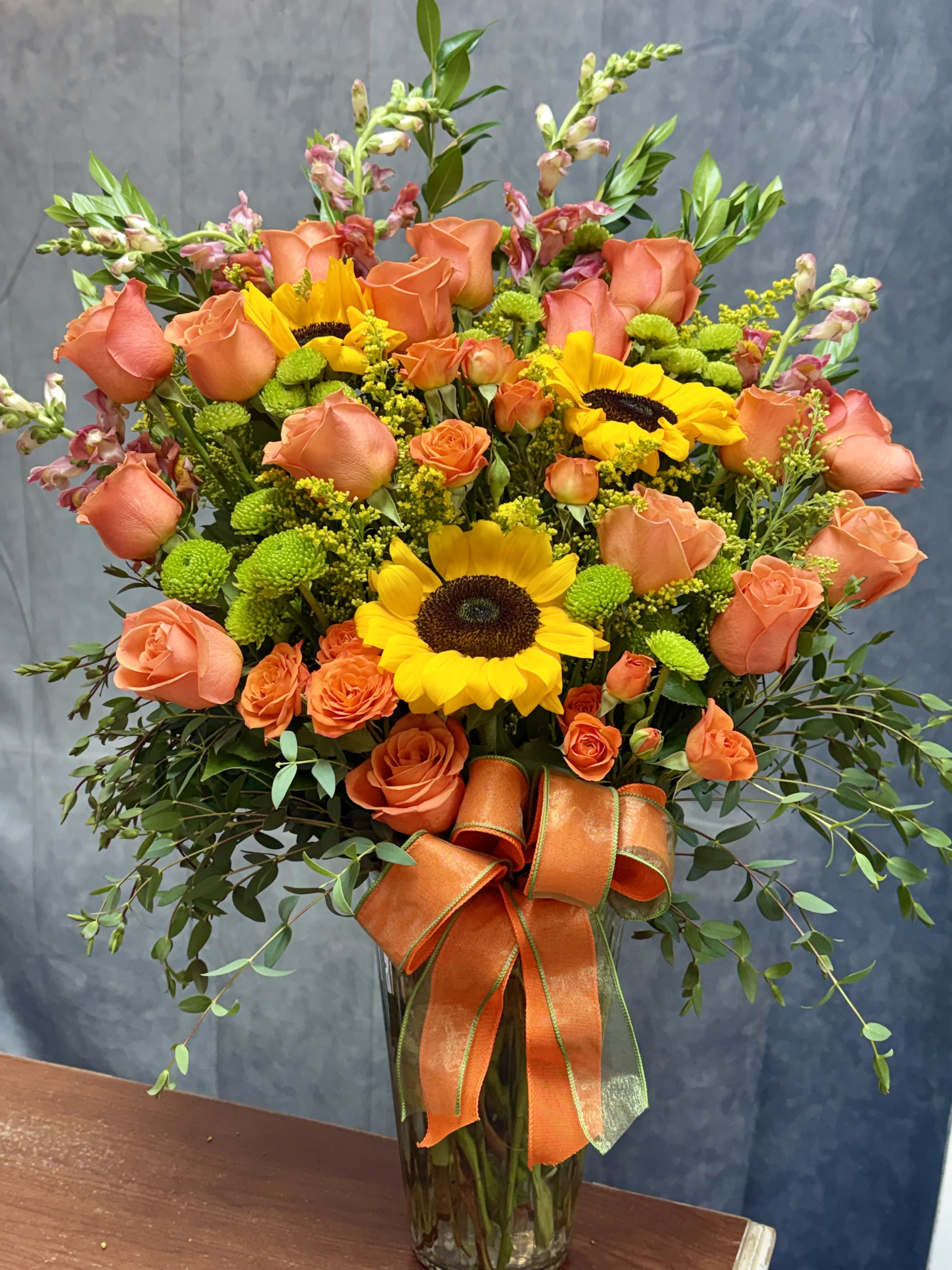 Bright Sunny Luxe Vase - A lush, oversized arrangement featuring sunflowers, premium roses, spray roses, snapdragons, and vibrant green button mums arranged in a classic glass vase.