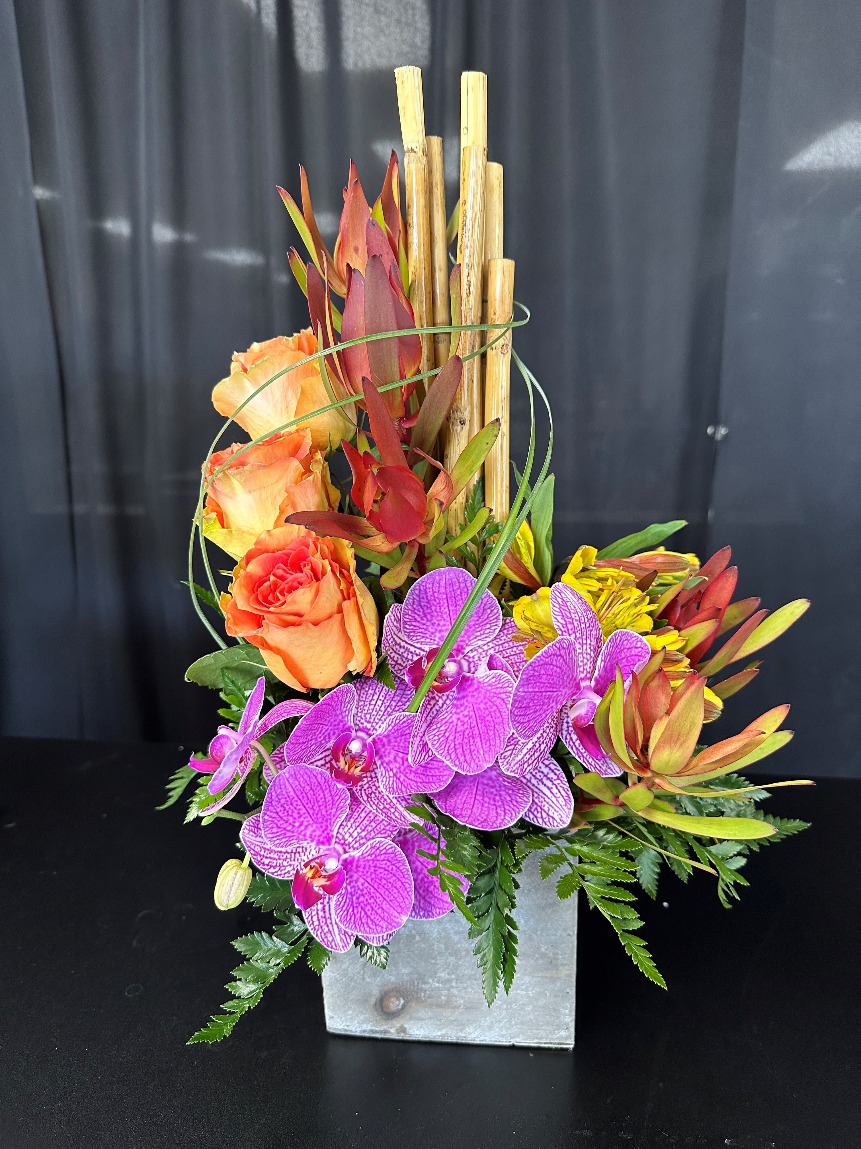 Orange roses and purple orchids in a square metal vase with bamboo accents