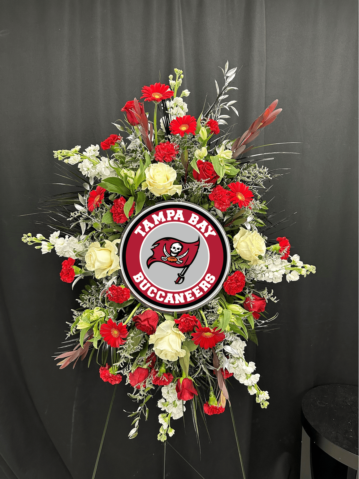 Custom Team Spray - Honor a devoted fan with a personalized standing spray designed in their favorite team’s colors. Our Custom Team Tribute features a vibrant mix of fresh seasonal blooms arranged around a centerpiece emblem that can be customized to reflect the team they loved most. From bold reds and whites to classic blues and golds, each design is thoughtfully created to capture team spirit while maintaining a dignified and elegant presentation.  Displayed on a standing easel, this tribute blends meaningful symbolism with heartfelt remembrance. Whether celebrating a lifelong Buccaneers fan or representing another beloved team, this spray offers a unique and personal way to honor their passion, loyalty, and legacy.  Team colors, ribbon accents, and centerpiece emblem are fully customizable.