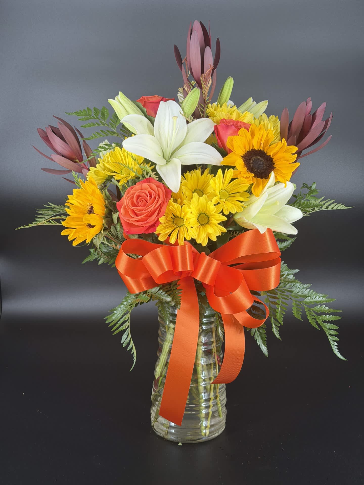 Mixed bouquet of lilies, sunflowers, roses, and yellow daisies in a glass vase with an orange ribbon