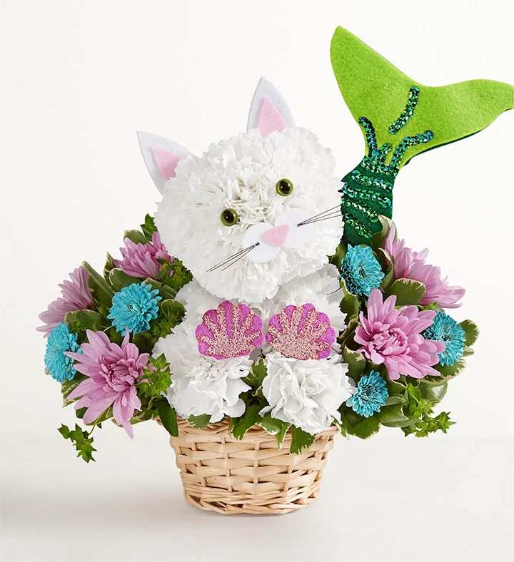 Seaside Mermaid™ Cat - EXCLUSIVE Our seaside-inspired mermaid kitty is making a splash for summer! Handcrafted with white carnations, she glitters in a lavender shell top and sequined green tail. Surrounded by cool-toned blooms, this coastal cutie is ready to send waves of happiness over birthdays, beach days… any day you want to make a little more magical.