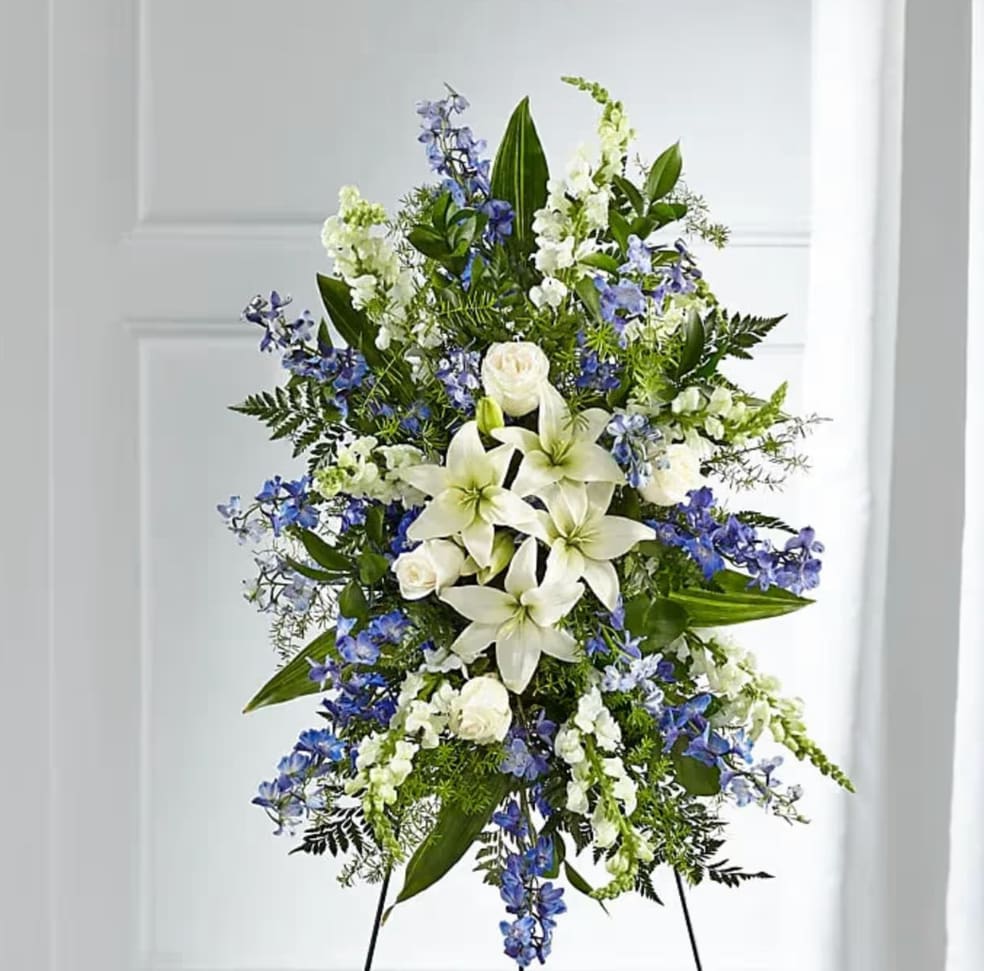 FAITHFUL  STANDING SPRAY - A graceful standing spray featuring an elegant blend of fresh white lilies, pure white roses, and striking blue delphinium. The serene white blooms symbolize peace, purity, and remembrance, while the soft blue accents add a touch of calm and reverence. Arranged with lush greenery on a standing easel, this tribute creates a dignified and soothing display, offering comfort and honoring a life with timeless beauty and heartfelt respect. 