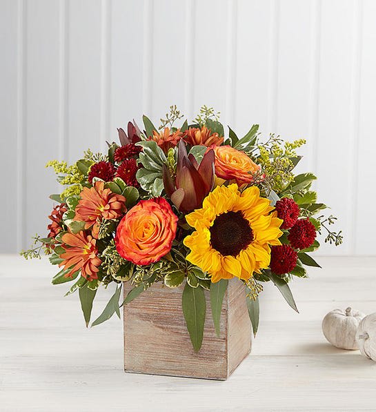 Harvest Glow Bouquet - The color and charm of an autumn country harvest inspired our farmhouse-style bouquet. A loose, natural gathering of blooms in vibrant shades of red, orange and yellow creates the perfect complement to our rustic, grey-washed wooden cube. With its soft tones and natural textures, it’s an ideal container for flowers and plants, adding warm and coziness to any fall décor.  All-around arrangement with red button poms and leucadendron, autumn-colored roses, sunflowers and bronze daisy poms; accented with red hypericum and assorted greenery Artistically designed in our exclusive grey washed wooden cube with rustic style and natural, textural beauty; measures 5.2"L x 5.2"H x 5.2"W Large arrangement measures approximately 12"H x 12"W Medium arrangement measures approximately 10"H x 10"W Small arrangement measures approximately 9"H x 9"W Our florists hand-design each arrangement, so colors and varieties may vary due to local availability Sunflowers may have light or dark centers
