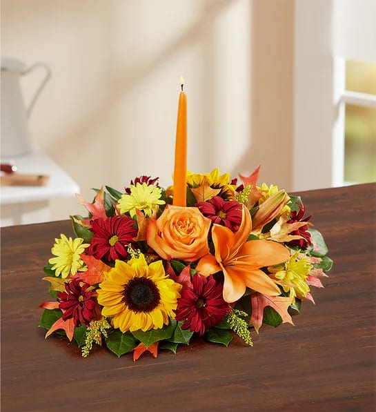 Fields of Europe Fall Centerpiece - The rich beauty of the European countryside comes alive in our best-selling fall centerpiece. Handcrafted by our artisan florists, it’s filled with a gathering of fresh-picked blooms in brilliant colors and finished with an orange candle to warm hearts and homes.  All-around centerpiece arrangement with autumn-colored roses, sunflowers, red daisy poms, orange Asiatic lilies, mini carnations and solidago; accented with assorted greenery and dried oak leaves