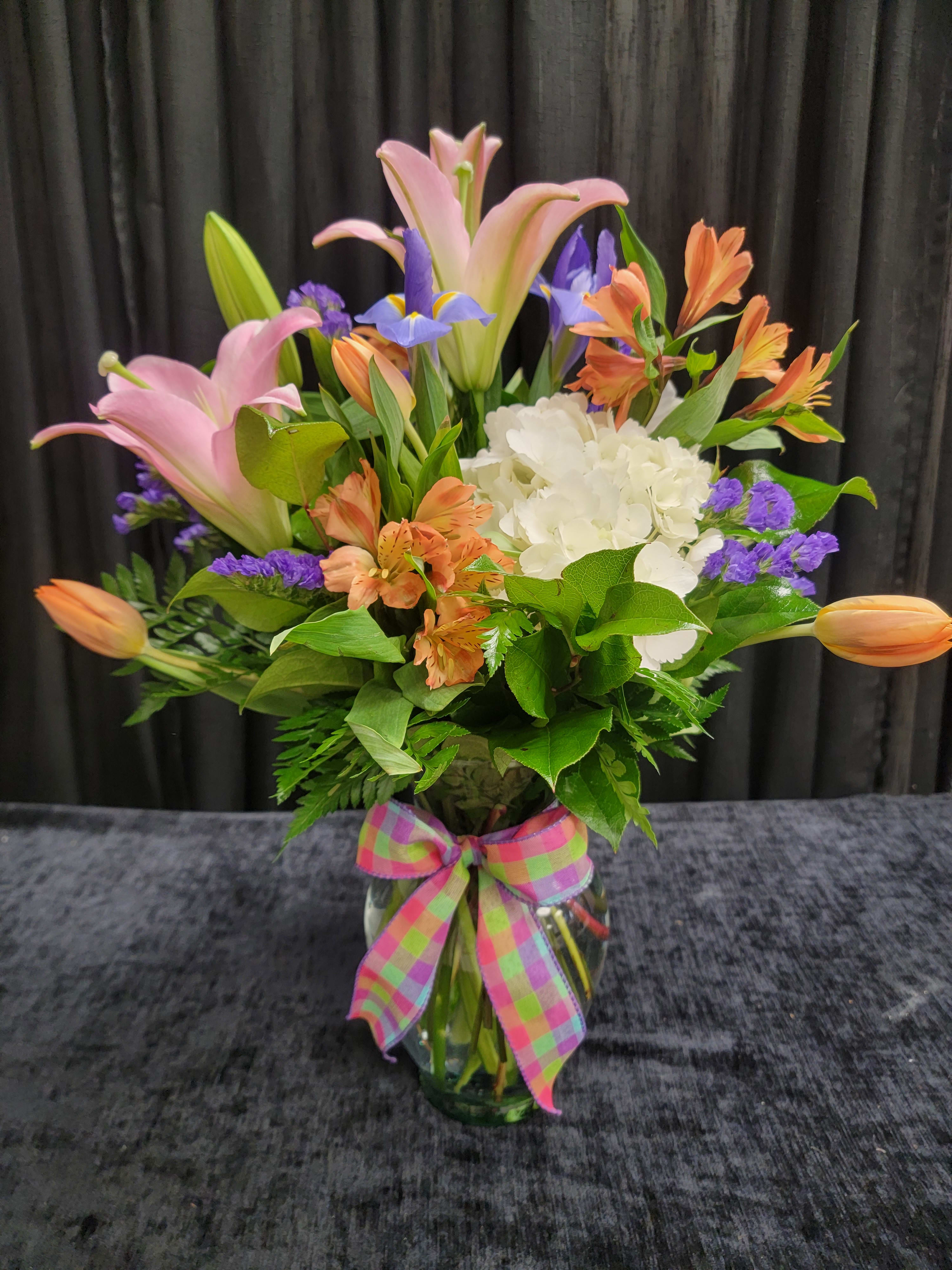 Mixed bouquet of pink lilies, orange blooms, white hydrangea and purple flowers in a glass vase with plaid bow.