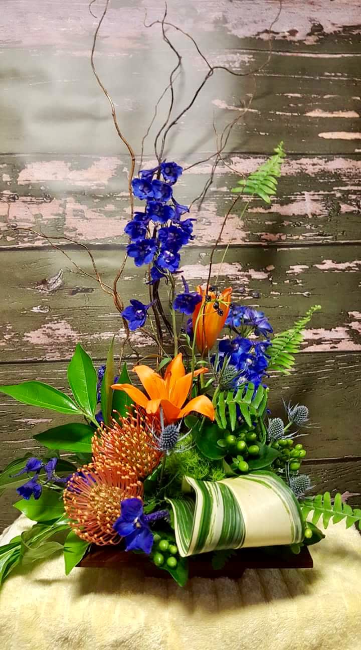 Tropical floral arrangement with orange lilies and blue flowers in a low container