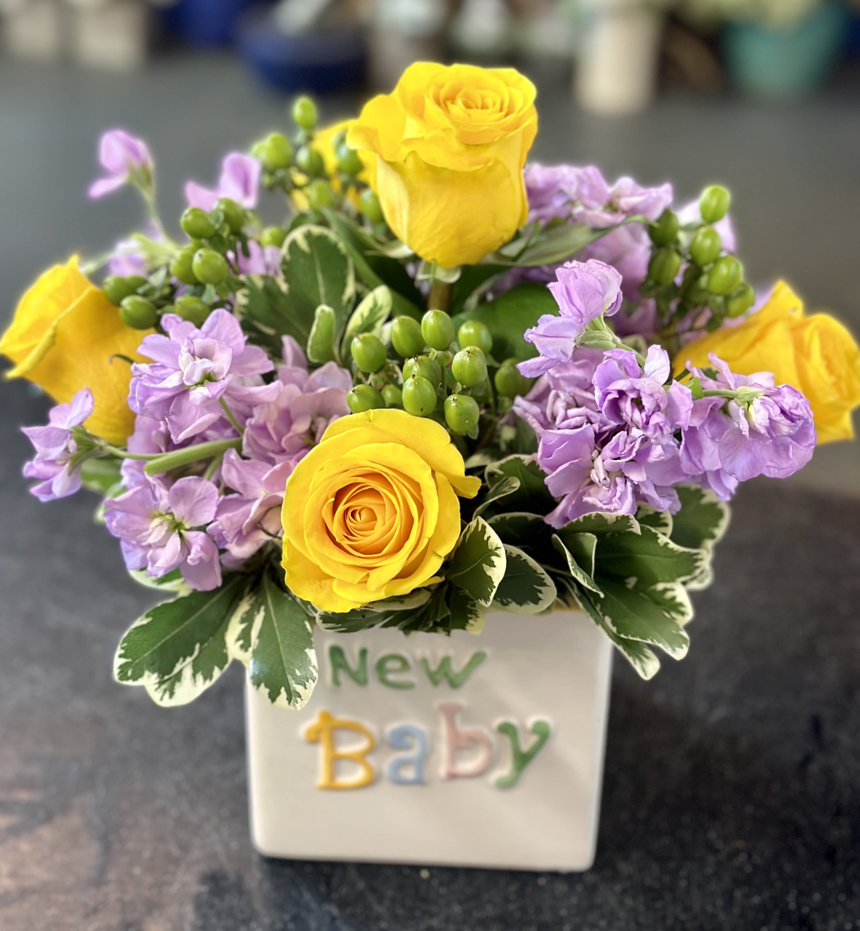 Yellow roses and lavender flowers in a white vase labeled New Baby