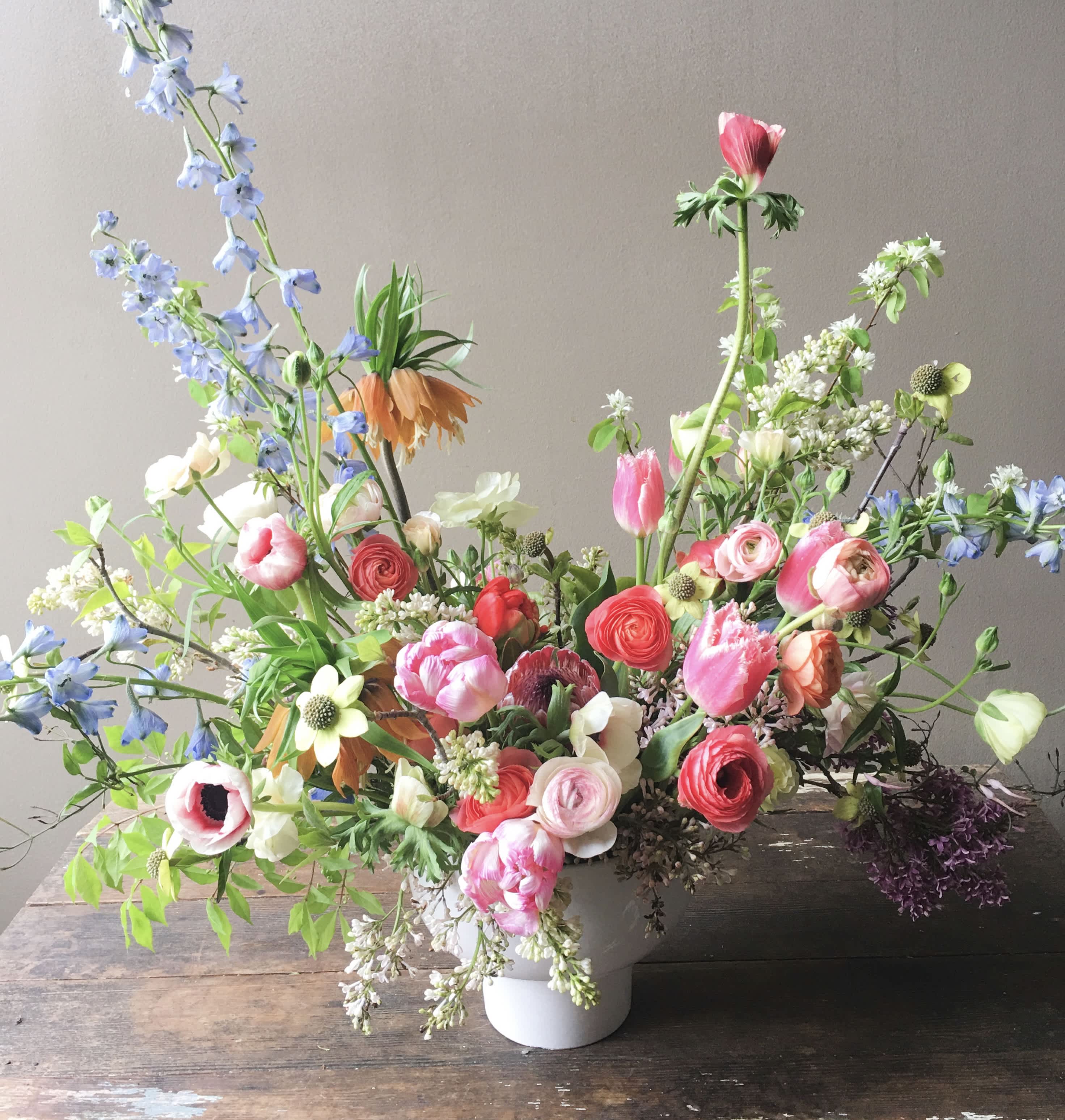 Asymmetrical garden-style arrangement of pink, coral, and white flowers with blue accents in a white vase