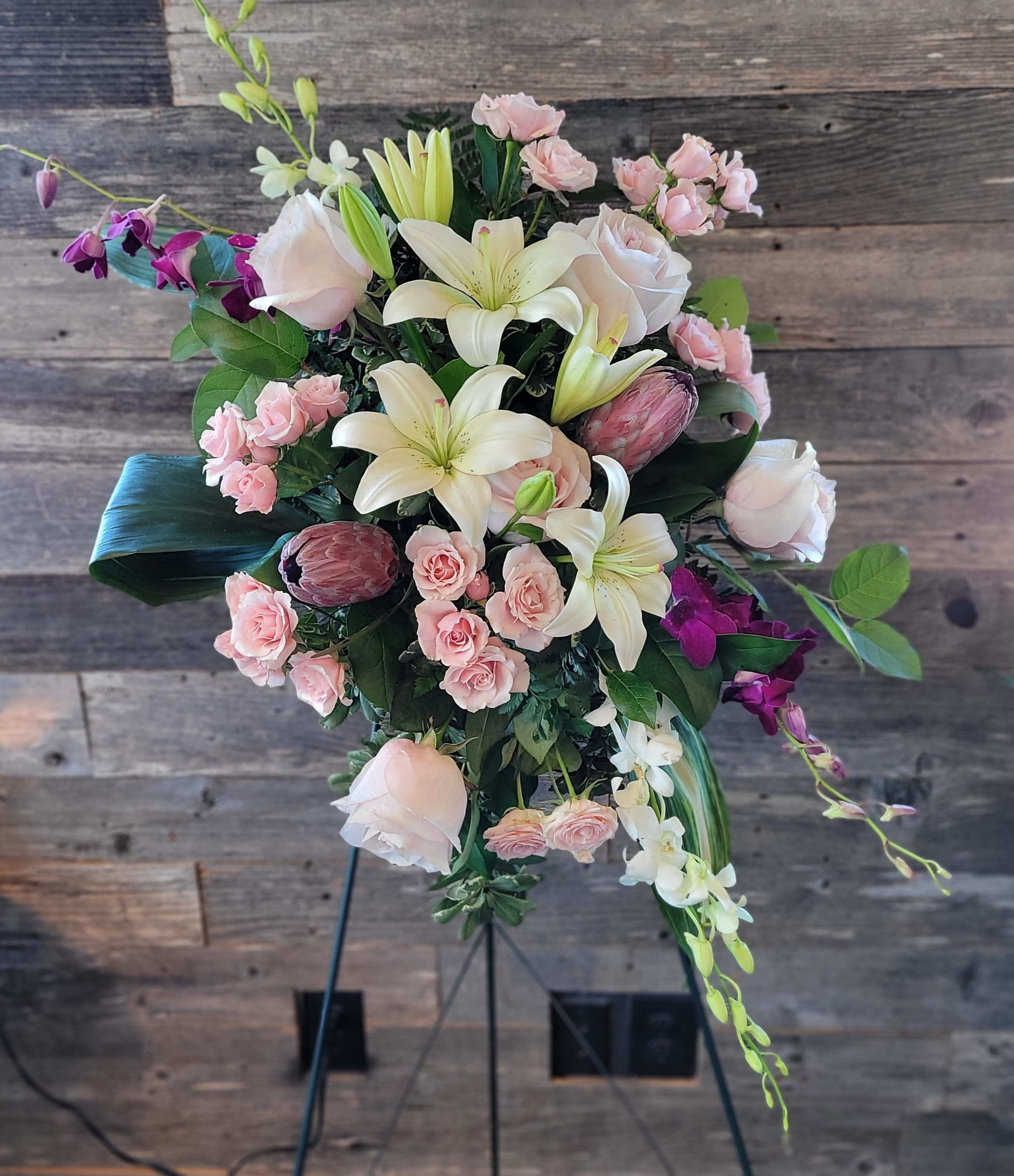 Tropical Standing Spray - Creamy white lilies spill from the center of this Tropical standing spray, their trumpet shapes and soft fragrance creating an immediate sense of graceful drama-perfect as an upscale sympathy and celebration display. Pale pink roses nestle among the lilies for a romantic focal point, while purple orchid sprays and spray roses add delicate contrast and texture that draw the eye along the cascading silhouette.  This large, cascading design sits on a sturdy floral stand and features trailing elements that give it a loose, flowing shape ideal for entryways. Protea adds an unexpected, sculptural touch amid the pink, white, purple, and green palette, making the arrangement both soft and striking. The scale makes it a statement piece for honoring someone special.
