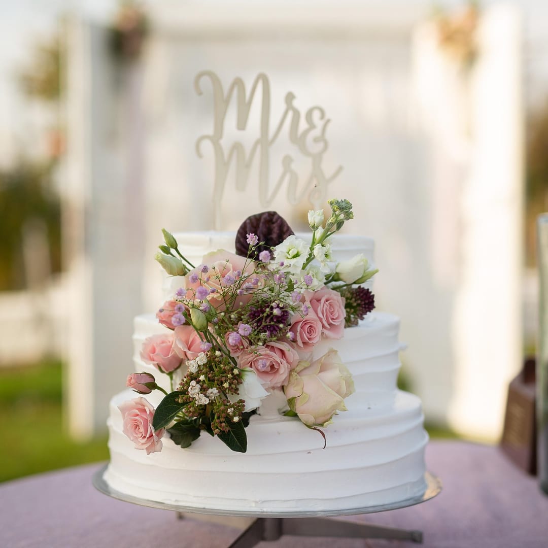 White tiered wedding cake topped with pink roses and small purple flowers