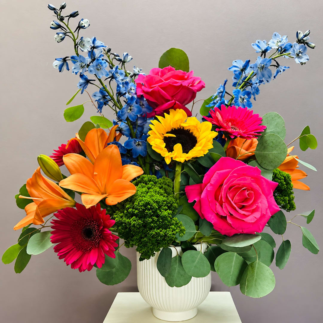Bright mixed bouquet in a white vase with pink roses, lilies, gerberas, and blue flowers