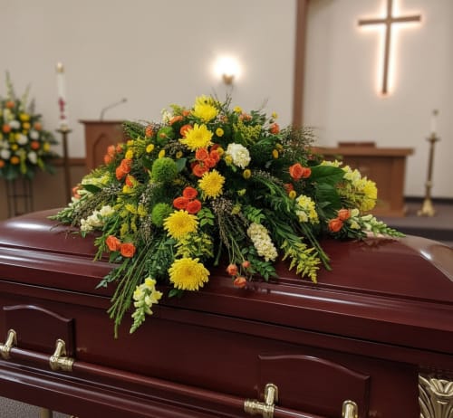 Casket spray of yellow, orange, and white flowers on a polished mahogany casket in a chapel.