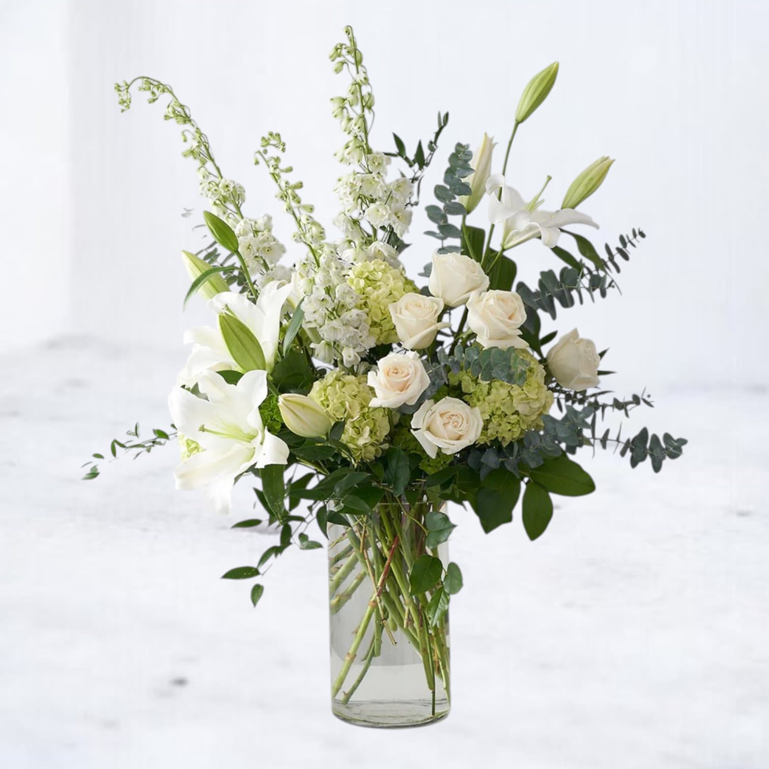 Sterling Passage - Sterling Passage is a graceful sympathy arrangement featuring elegant white lilies, soft ivory roses, hydrangea, and delicate delphinium arranged among lush seasonal greenery. The airy composition and soft white palette create a peaceful and refined floral tribute.  Designed in a tall glass vase with natural movement and layered textures, this arrangement offers a beautiful expression of comfort, remembrance, and support.  Available for same-day flower delivery in Magnolia, Montgomery, The Woodlands, Conroe, and surrounding Houston areas. Flower varieties may vary slightly based on seasonal availability while maintaining the soft white palette and overall design style.