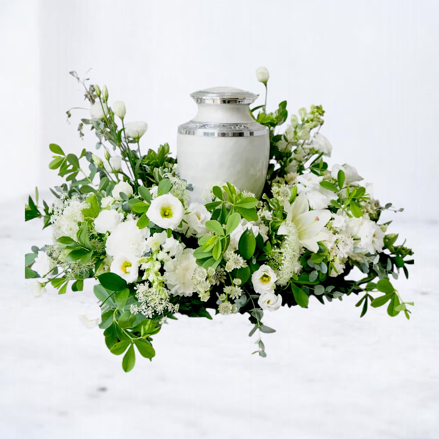 Palladium - Palladium is a modern white urn surround designed with lush seasonal greenery, white lisianthus, hydrangea, premium roses, delicate textural blooms, and soft garden florals surrounding a cremation urn in an airy asymmetrical composition. Designed with movement, softness, and layered texture, this arrangement creates a contemporary memorial display that feels peaceful, refined, and deeply elegant.  Featuring a fresh palette of crisp white florals and natural greenery, Palladium offers a modern alternative to traditional sympathy flowers while still honoring the beauty and significance of remembrance. Its sculptural design style makes it ideal for celebration of life services, memorial gatherings, funeral displays, or private home tributes.  Handcrafted with premium seasonal flowers and available for same-day sympathy flower delivery in Magnolia, TX and surrounding areas including The Woodlands, Montgomery, Conroe, Spring, Tomball, and nearby Lake Conroe communities.