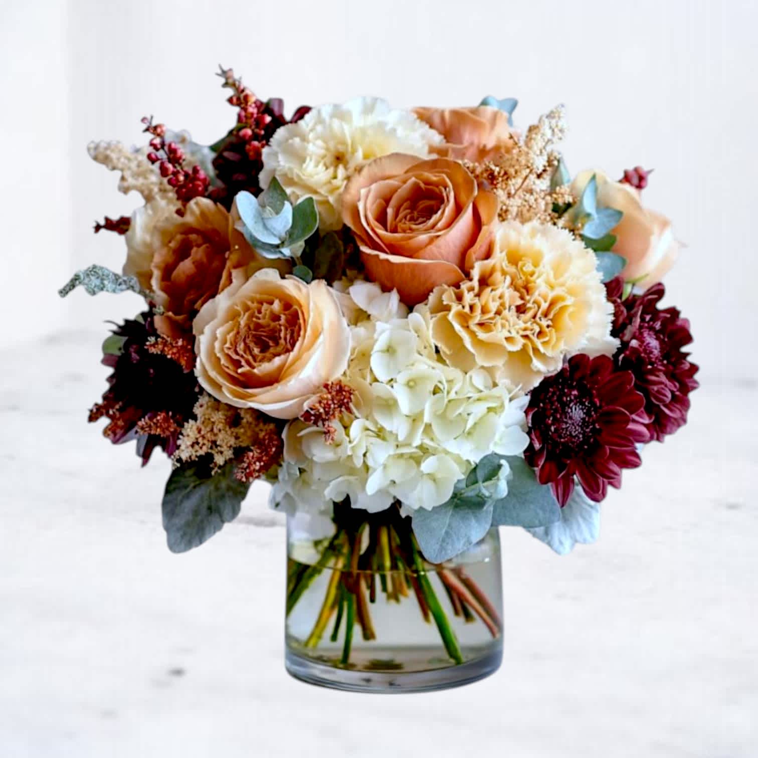 Bouquet of peach roses, white hydrangeas, and burgundy blooms in a glass vase