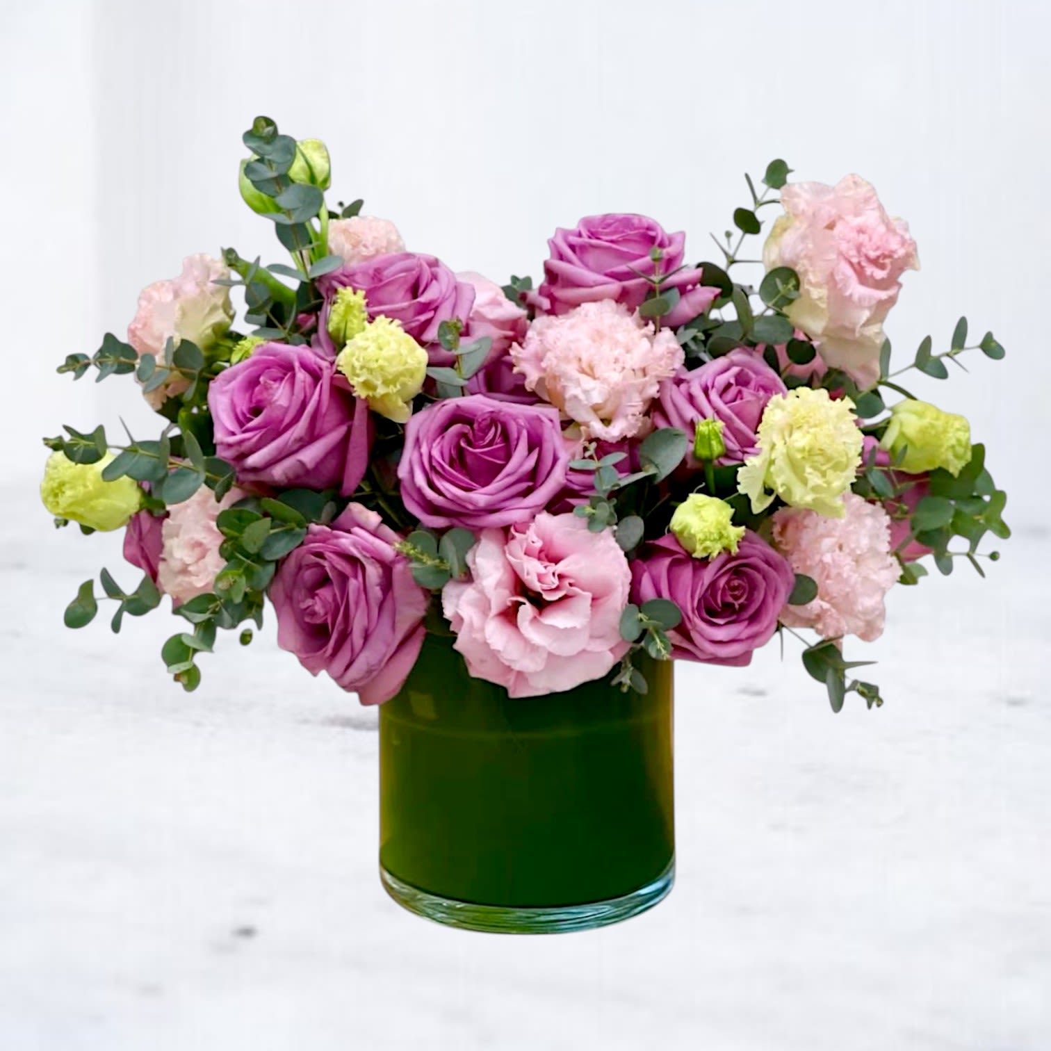 Lavender Harmony - A soft, romantic cluster of lavender and pink roses with pale pink lisianthus and light green carnations - Lavender Harmony is a low, rounded flower arrangement perfect for anniversary bouquets, birthdays, or a "thinking of you" surprise. The palette of lavender, pink, purple, green, white and soft yellow reads both fresh and feminine, making this a lovely choice for love and romance, a new baby celebration, or a heartfelt "I'm sorry."  The blooms are densely arranged into a full, rounded shape with airy eucalyptus accents that add movement and contrast against the layered petals. Presented in a polished green glass vase, the composition feels contemporary yet quietly elegant - great for home décor, a desk surprise for Boss's Day, or a bedside table after a hospital visit. Sized for easy display, Lavender Harmony offers same-day delivery in Houston, Magnolia, Tomball, Cypress and surrounding areas for last-minute birthdays or thoughtful, just-because moments.
