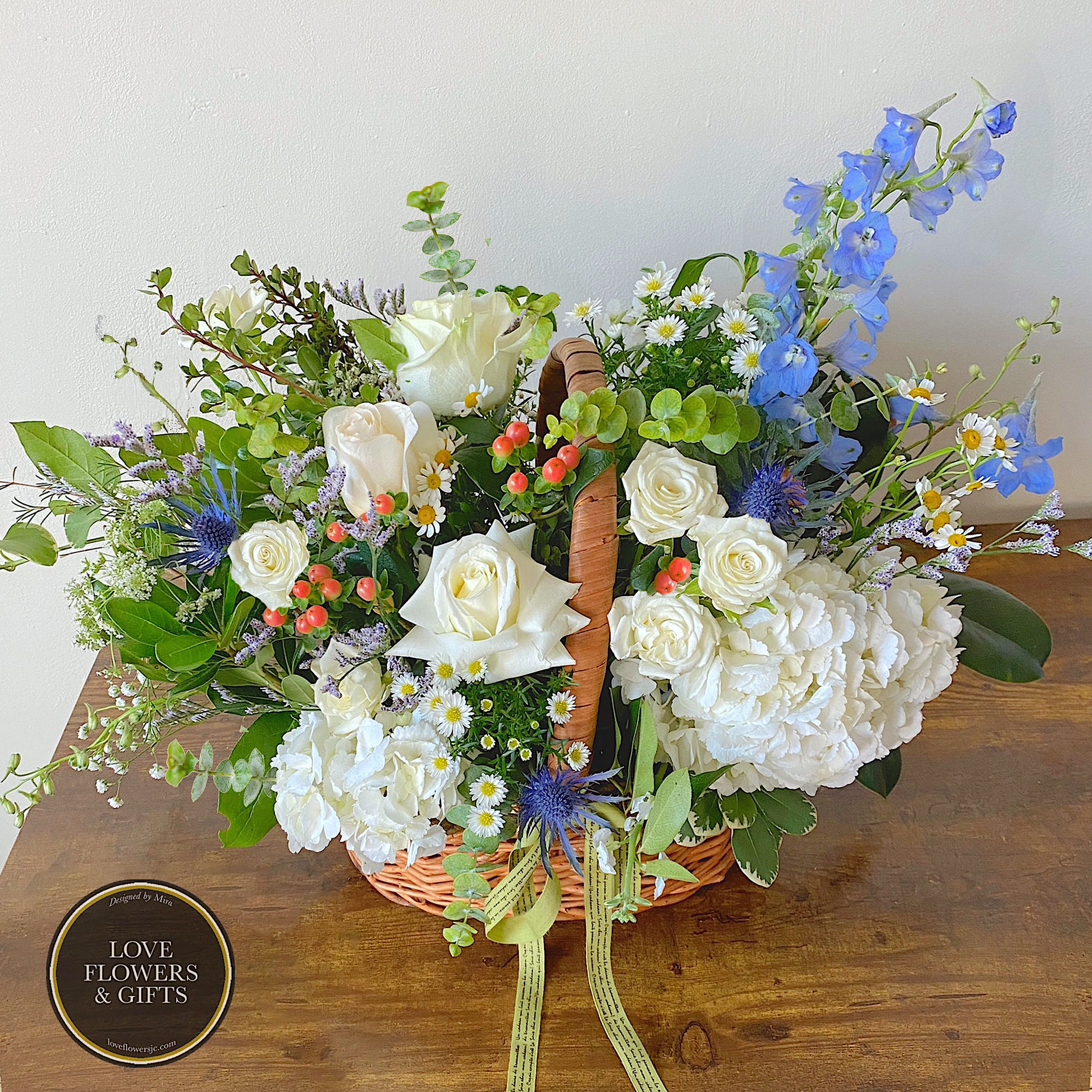 Basket arrangement of white roses, hydrangeas, and blue flowers with berries and a ribbon.